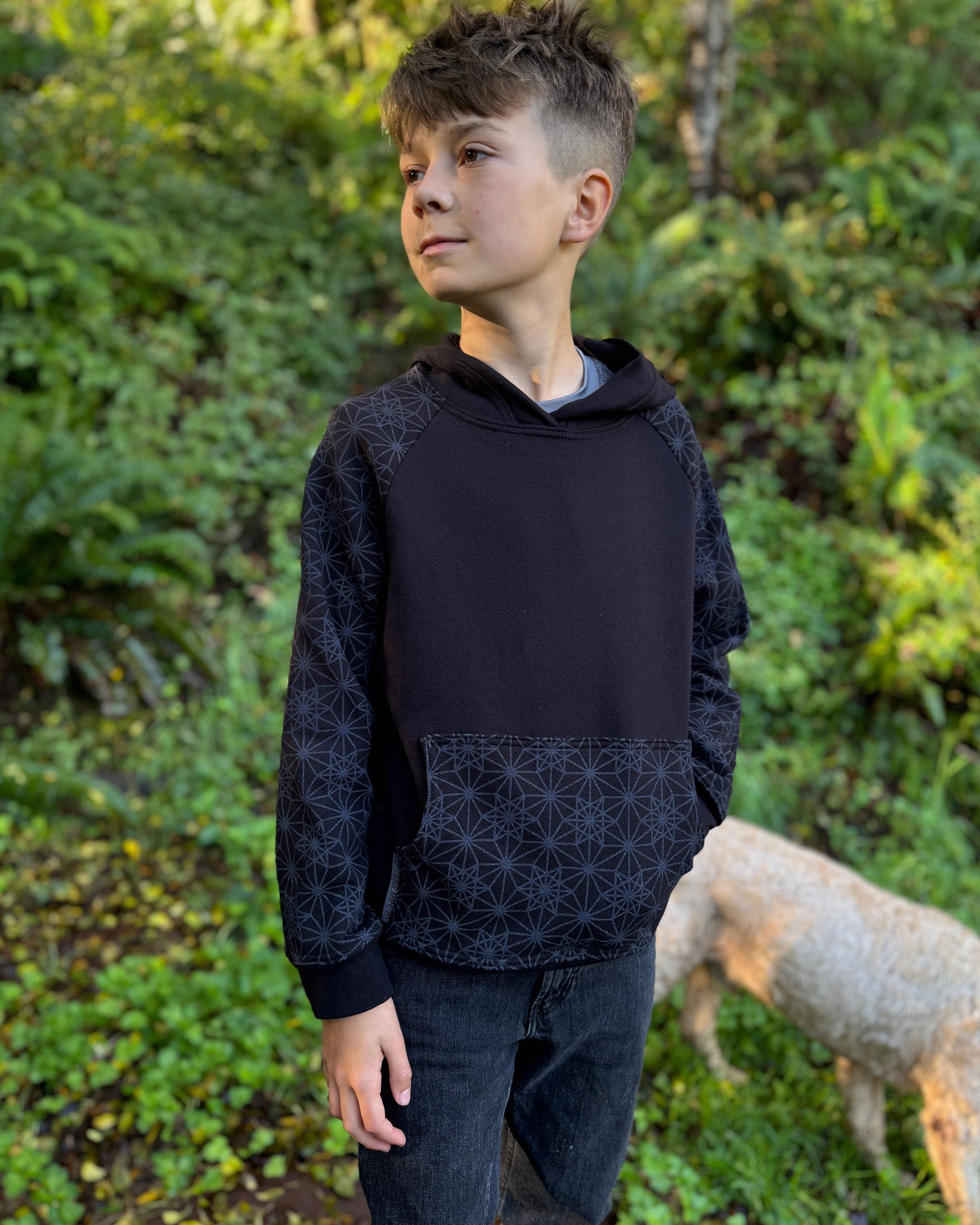 Organic cotton youth fleece hoodie, black with geometric printed sleeves, front right view, worn by a boy in nature.