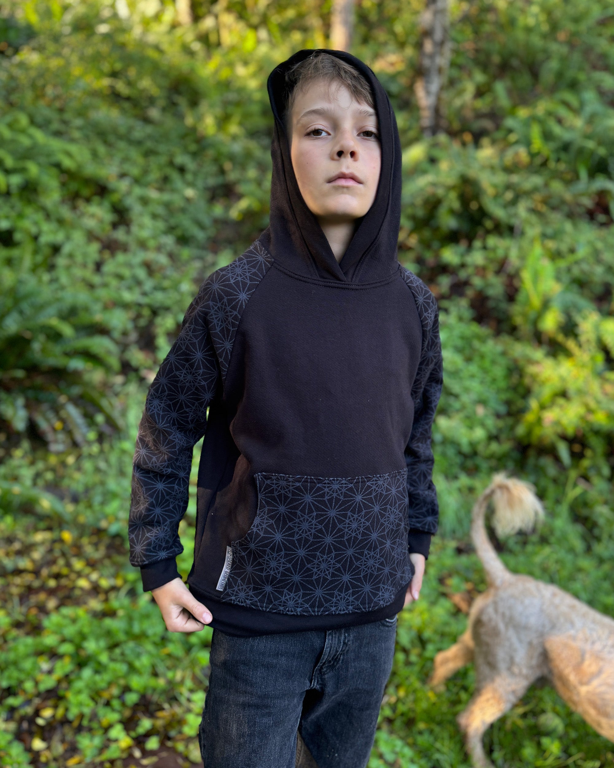 Organic cotton youth fleece hoodie, black with geometric printed sleeves, front right view, worn with hood up by a boy in nature.
