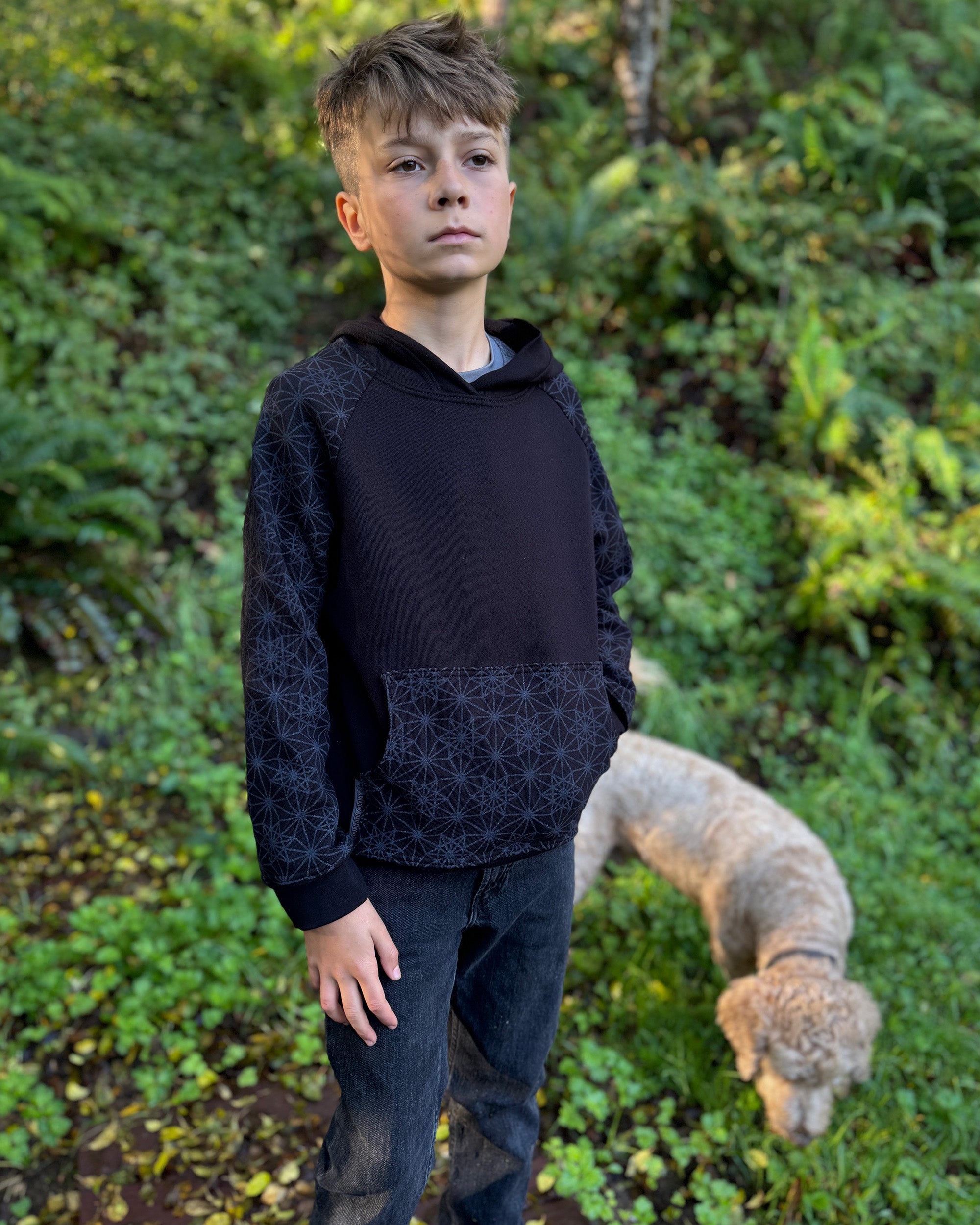 Organic cotton youth fleece hoodie, black with geometric printed sleeves, front left view, worn by a boy in nature.