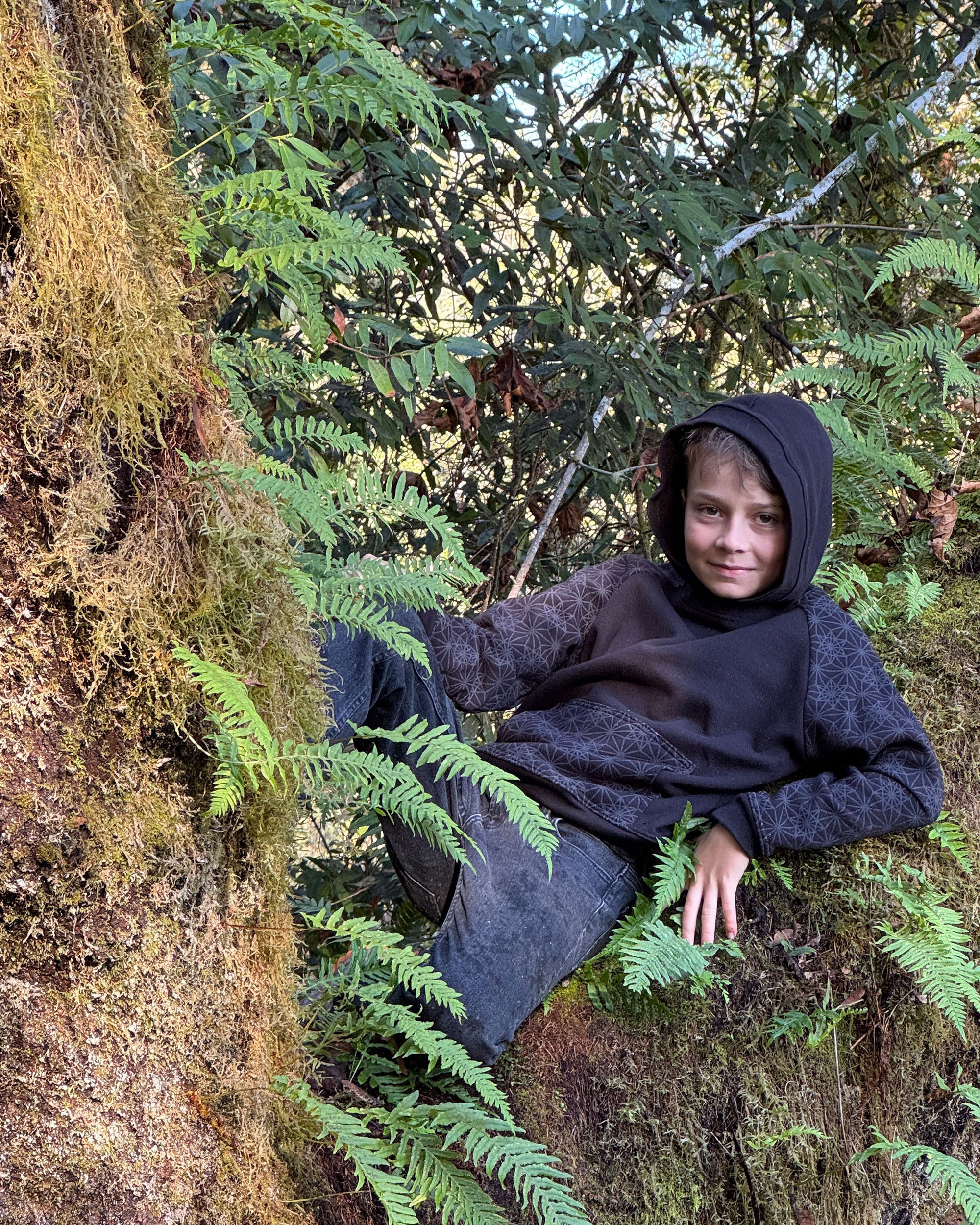 Young boy sitting among ferns and trees in a forest setting wearing a black geometric hoodie.