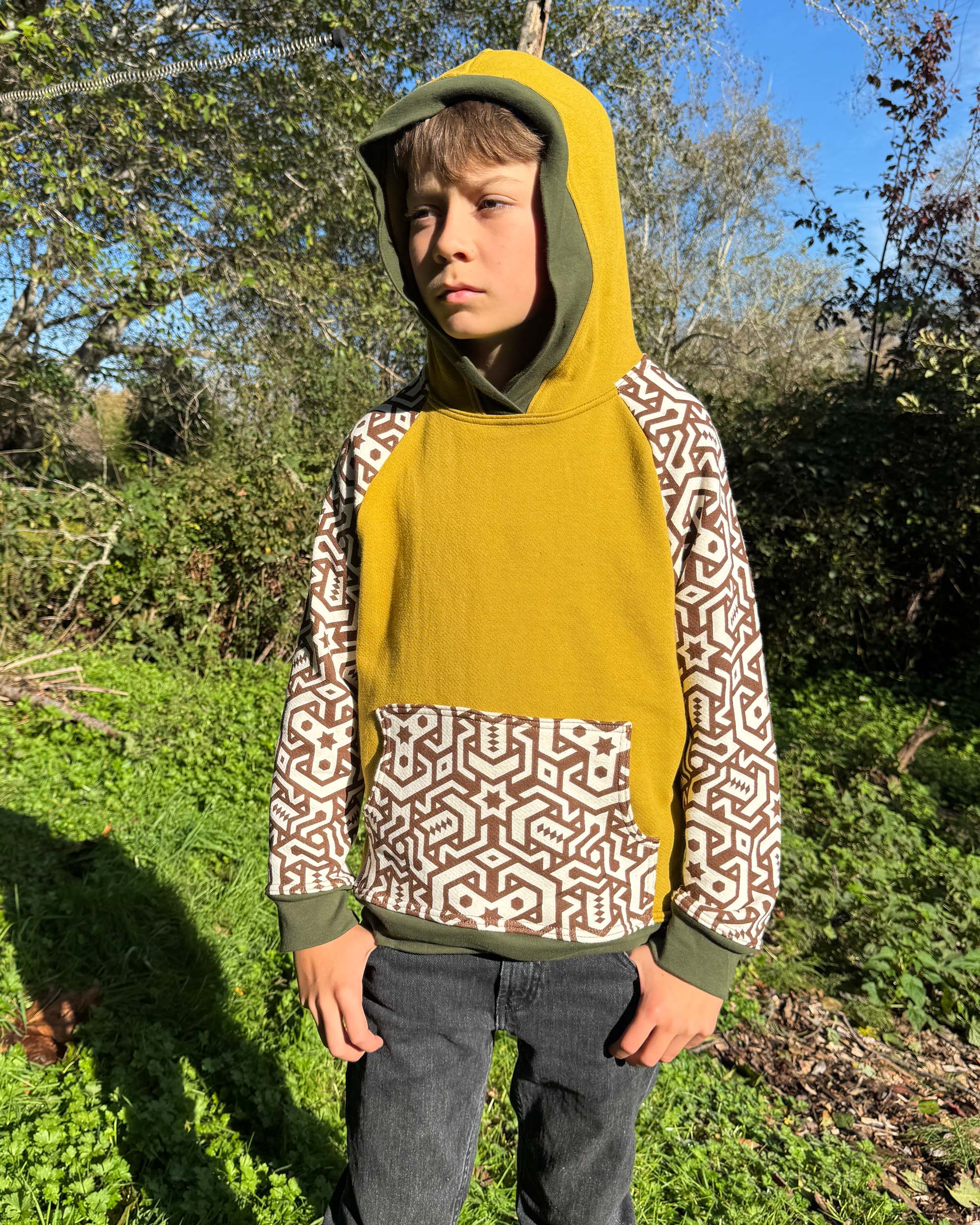 Organic cotton kids hoodie, yellow with geometric printed sleeves, front view, worn with hood up by a boy in nature.
