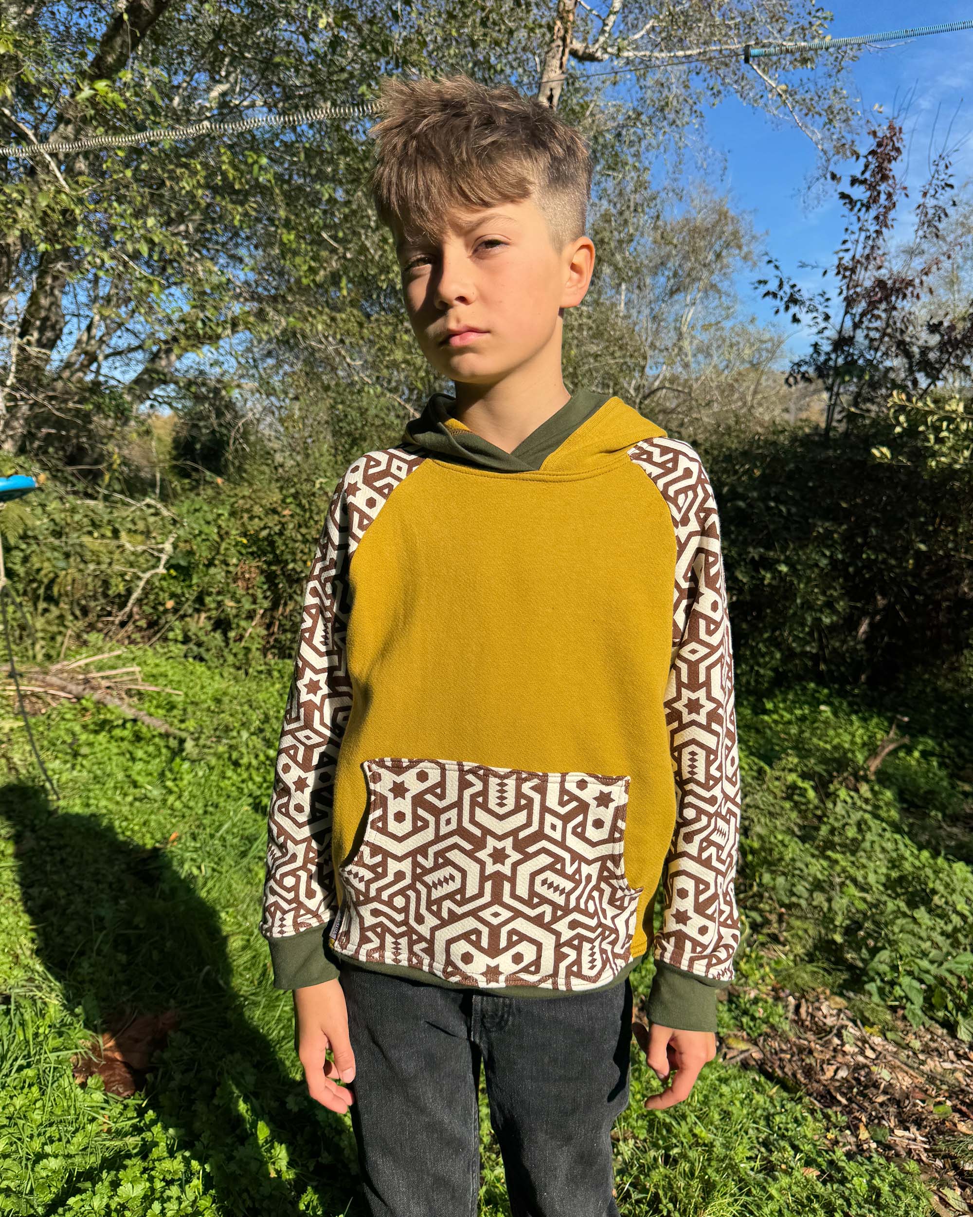 Organic cotton kids hoodie, yellow with geometric printed sleeves, front view, worn by a boy in nature.