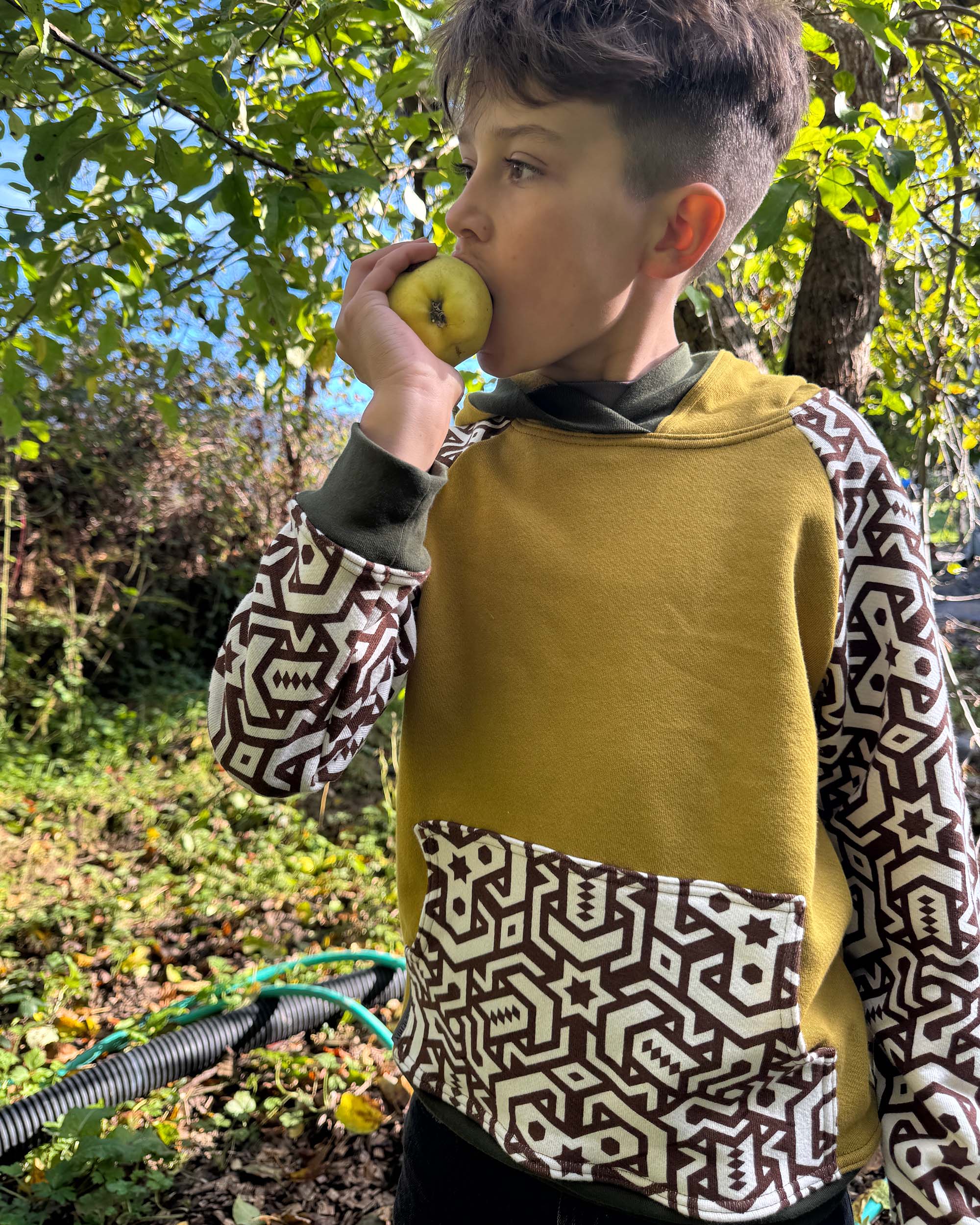 Boy eating an apple outdoors wearing yellow and geometric printed organic cotton hoodie.