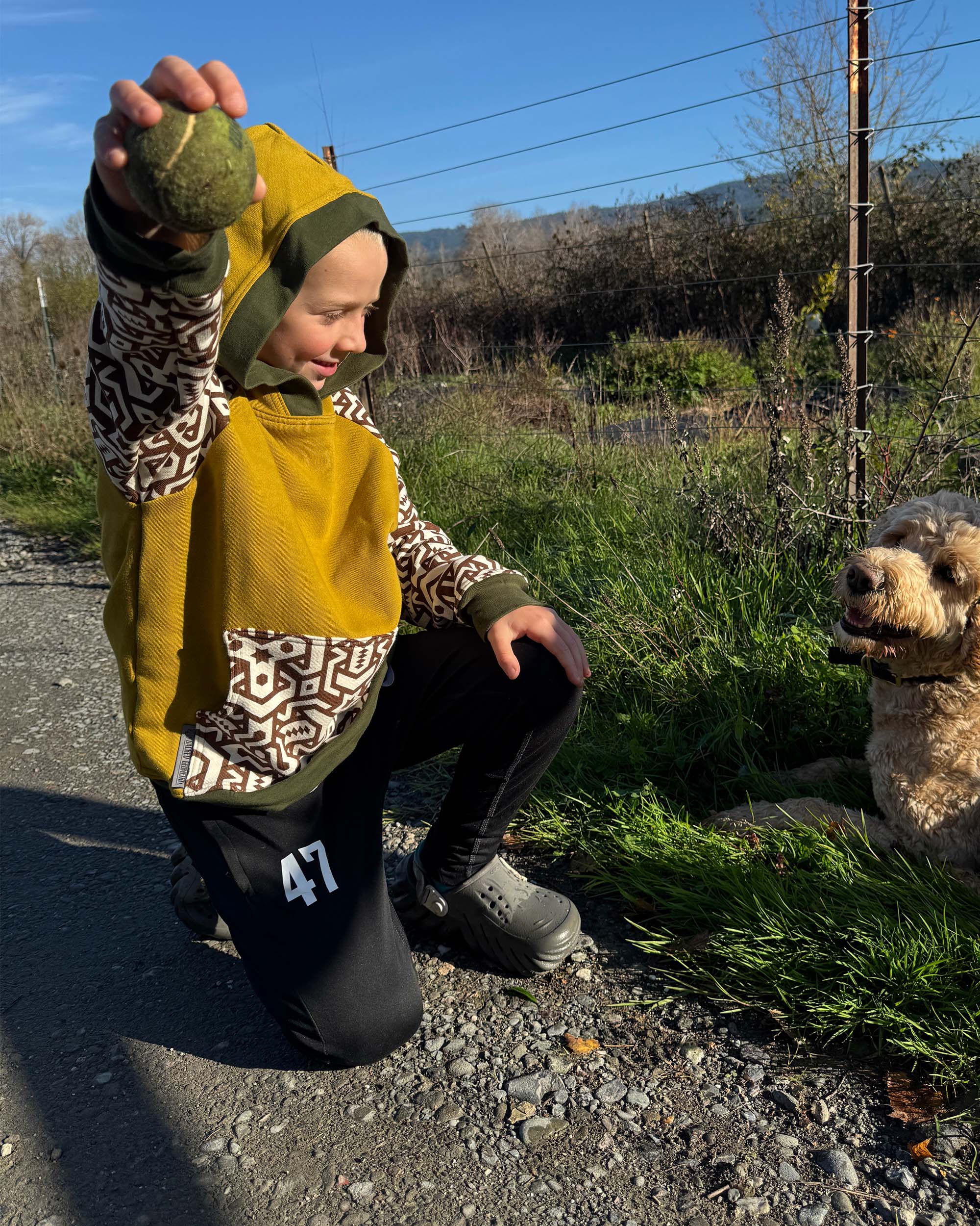 Child in a yellow sacred geometrix hoodie playing with his dog near a fence.