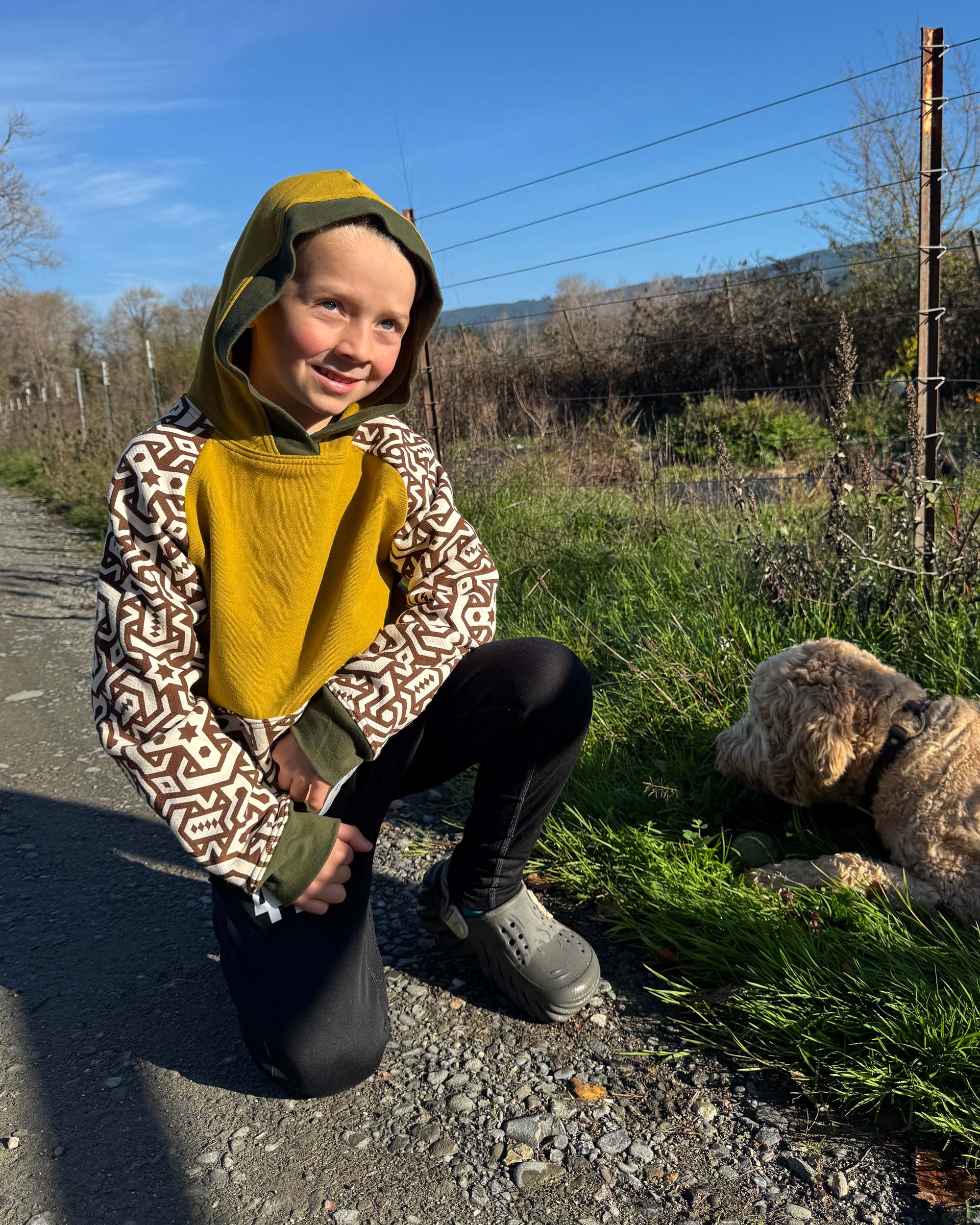 Organic cotton kids hoodie, yellow with geometric printed sleeves, front view, worn by a crouching boy with his dog.
