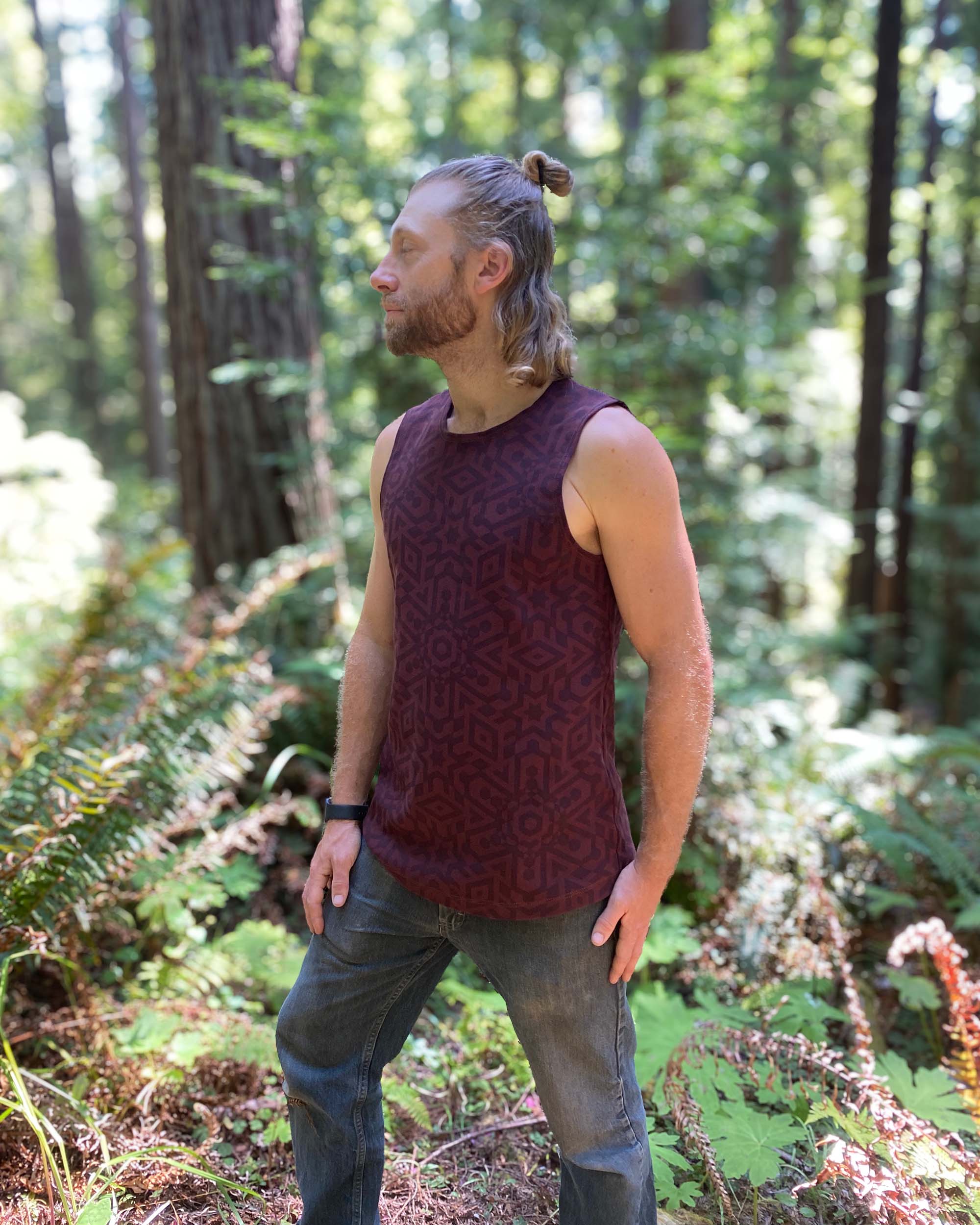 Organic cotton mens tank top, brown fabric with geometric print, right side view with model in nature and trees.