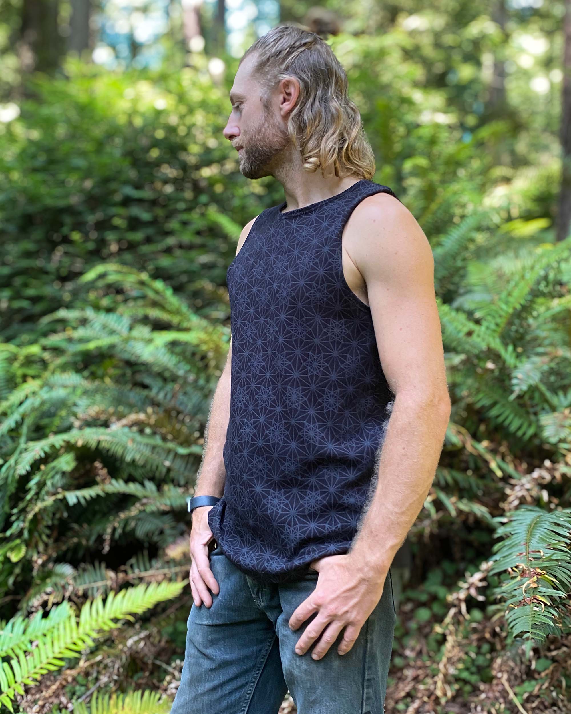 Organic cotton mens tank top, black fabric with geometric print, left side view with model in natural light.