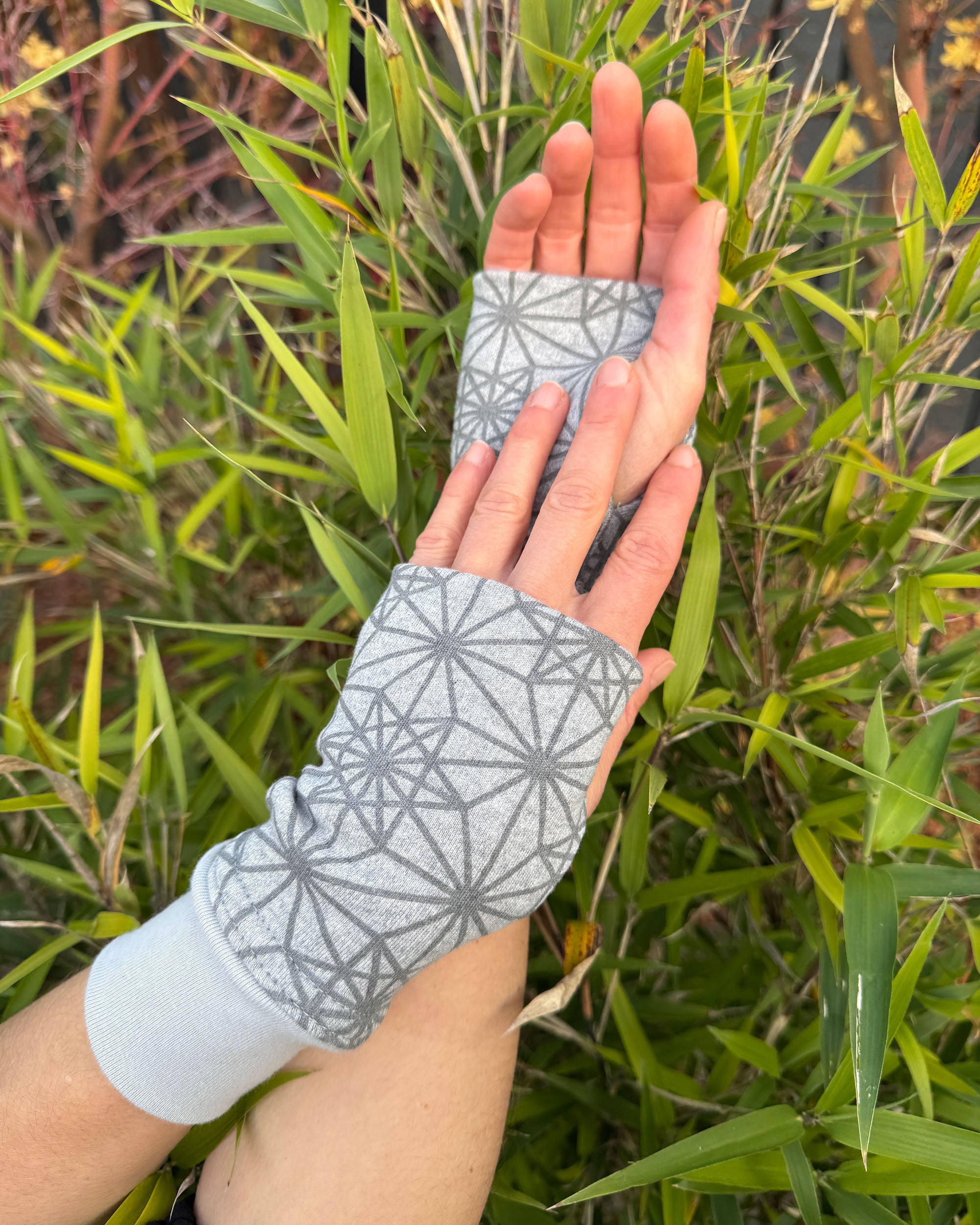 Hands wearing grey colored geometric organic cotton hand warmers, with nature background.