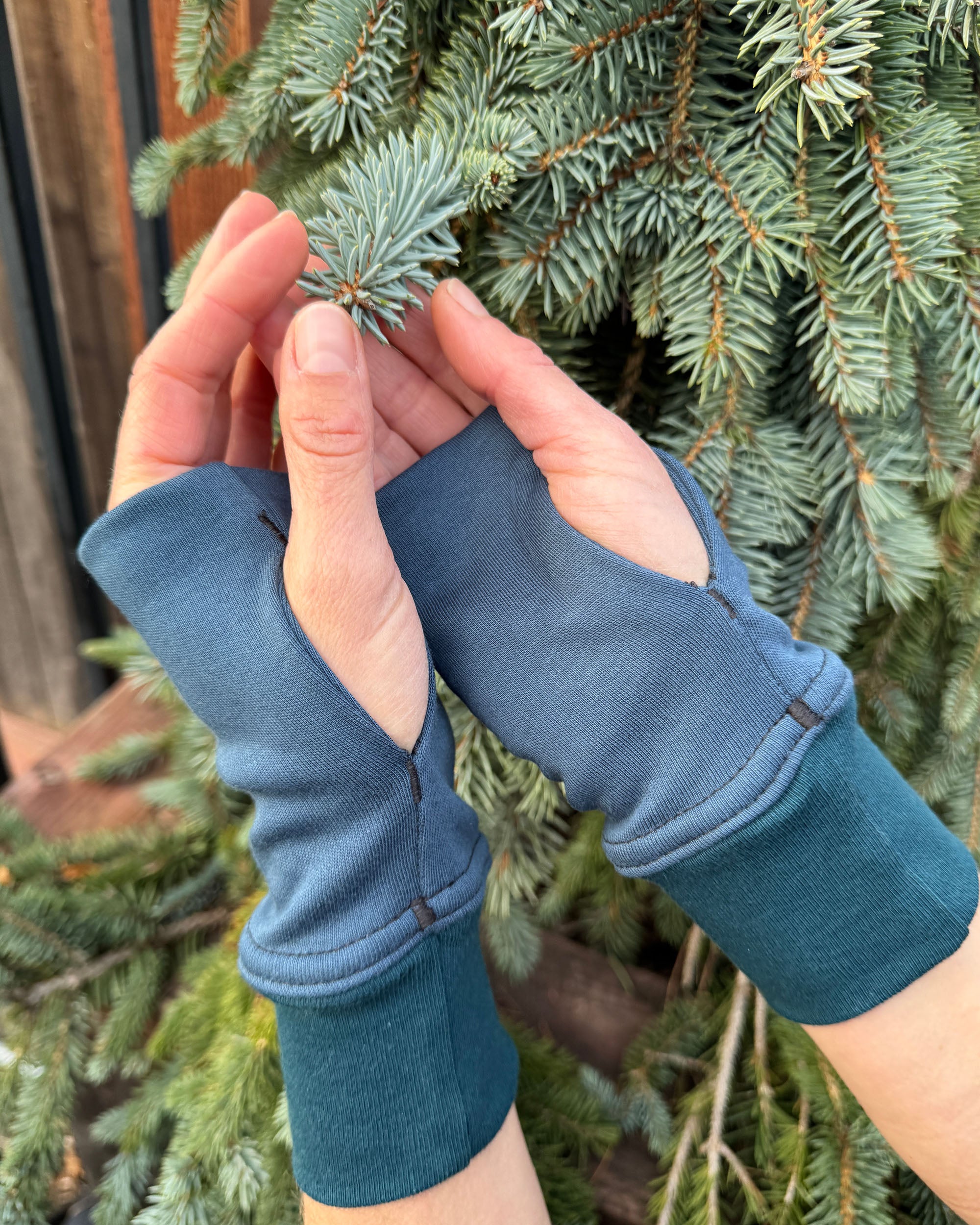 Female hands wearing dark blue organic cotton hand warmers shown with leafy background.