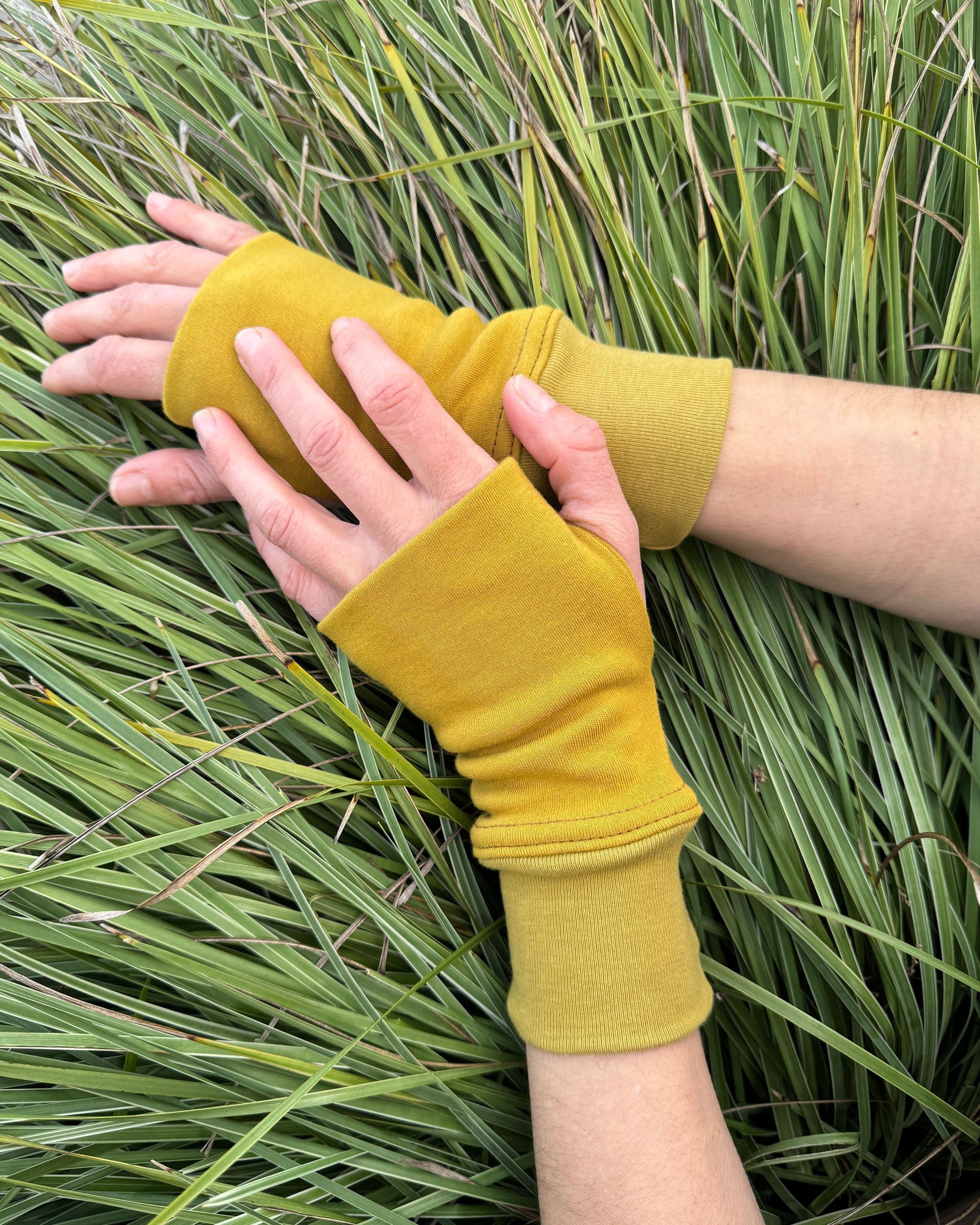 Female hands wearing organic cotton wrist warmers, yellow solid fabric with grassy background.