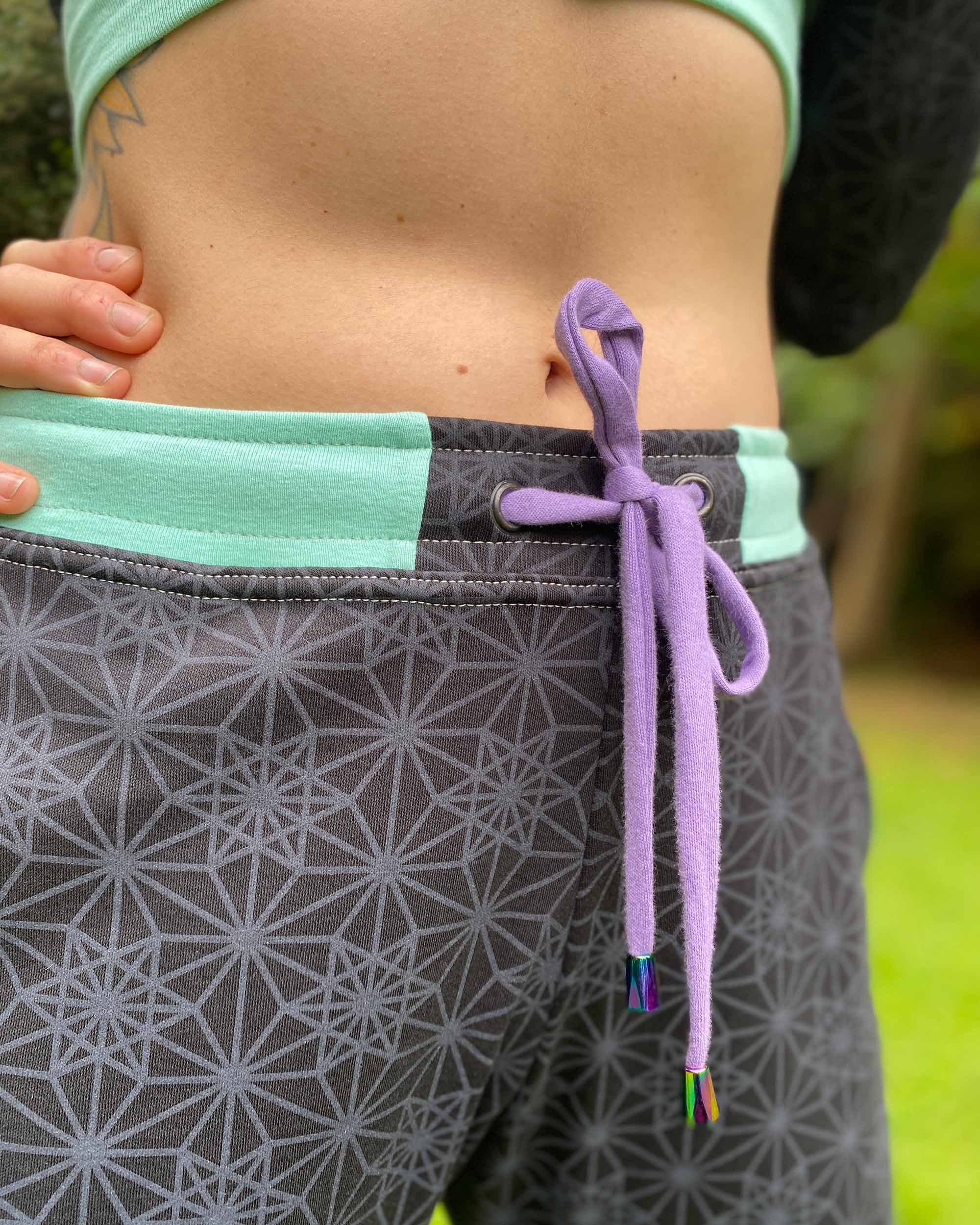 Organic cotton unisex joggers, black fabric with geometric print and purple accents, close up view of drawcord.