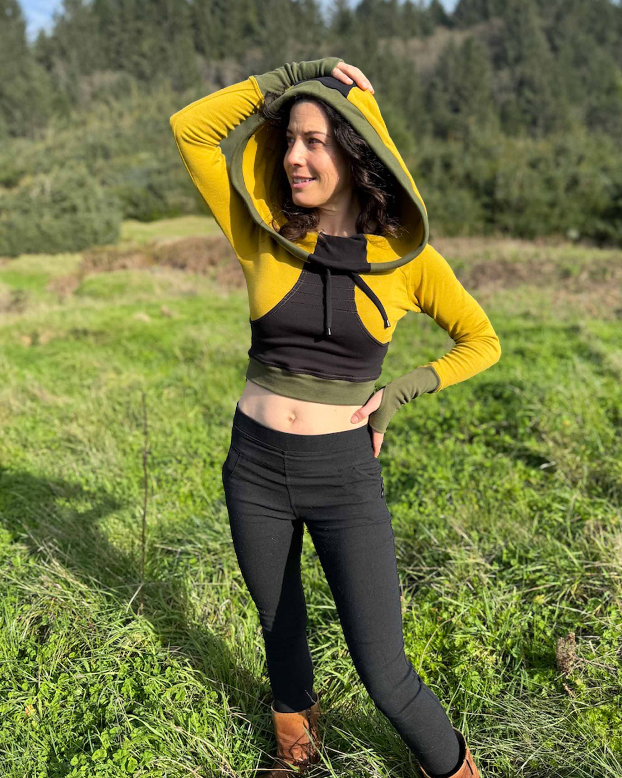Fitted crop hoodie with cowl hood, yellow fabric with black accent, front side view on model in grass.