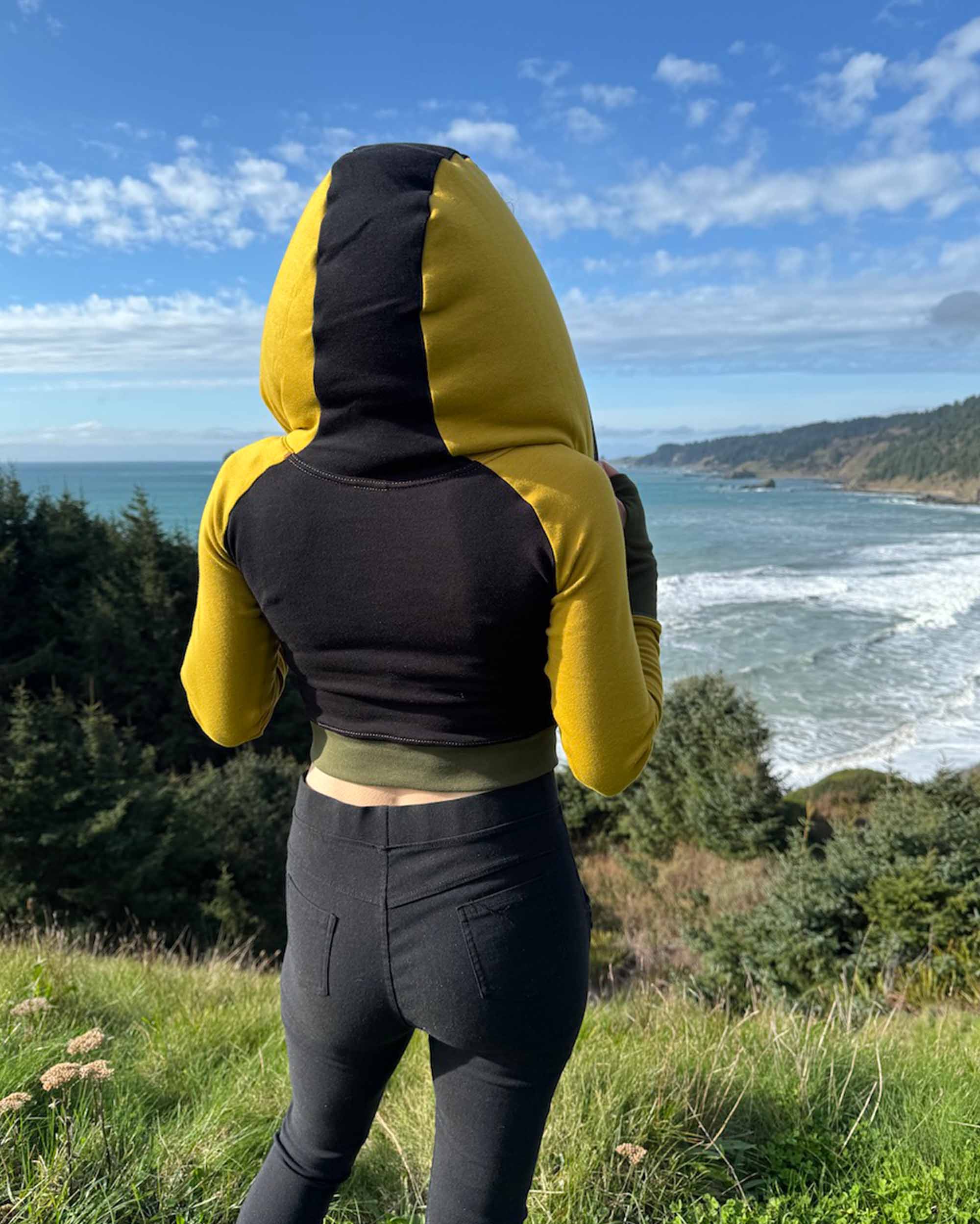 Fitted crop hoodie with cowl hood, yellow fabric with black accent, back side view on model.
