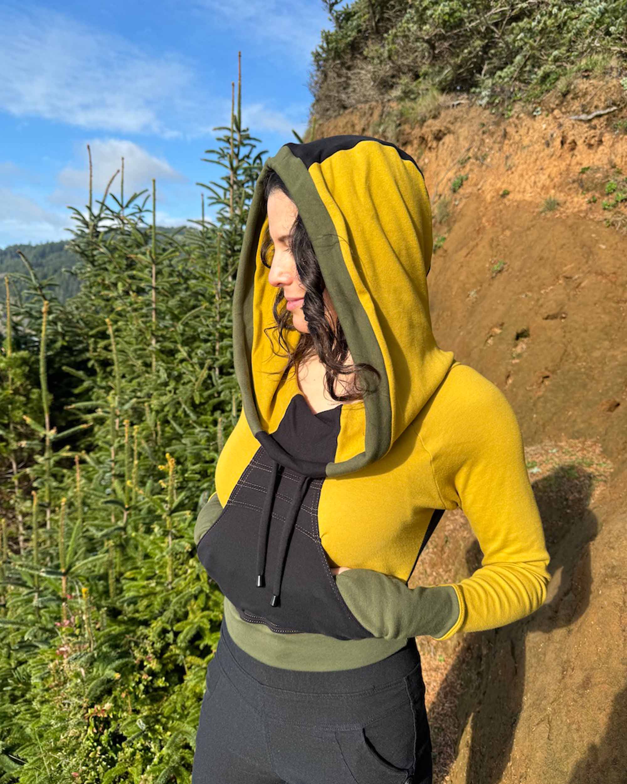 Fitted crop hoodie with cowl hood, yellow fabric with black accent, left side view on model in trees.