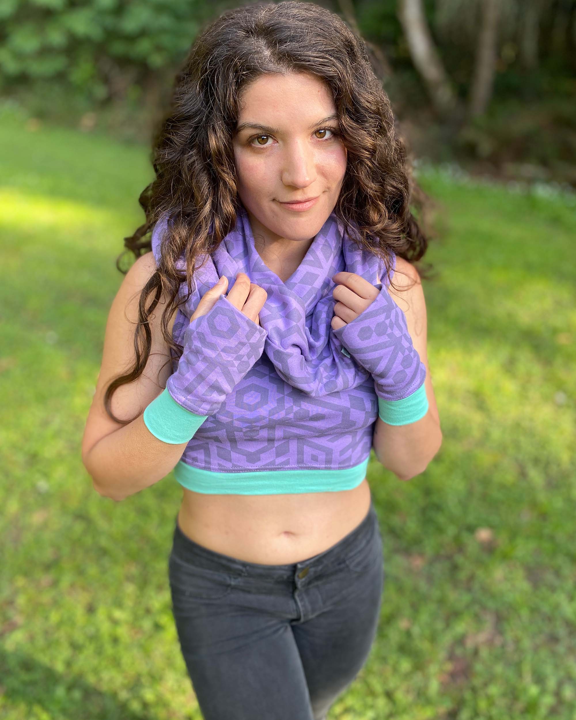 Female model wearing matching set of crop top, infinity scarf and fingerless gloves, lavender organic cotton fabric with geometric print, front view in grass field.