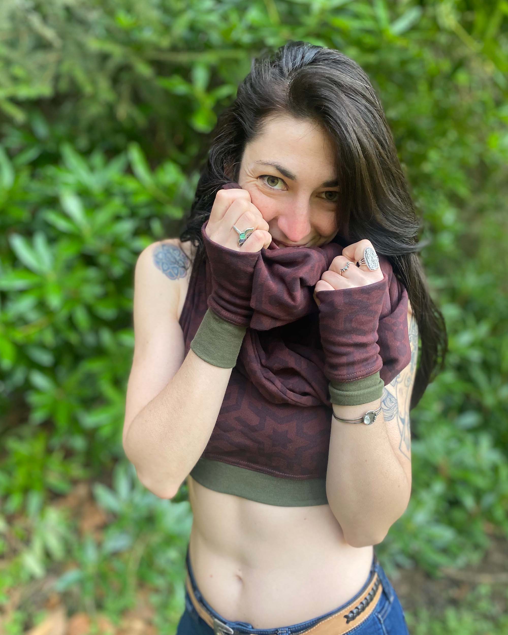 Female model wearing matching set of crop top, infinity scarf and fingerless gloves, brown organic cotton fabric with geometric print, front view in natural setting.