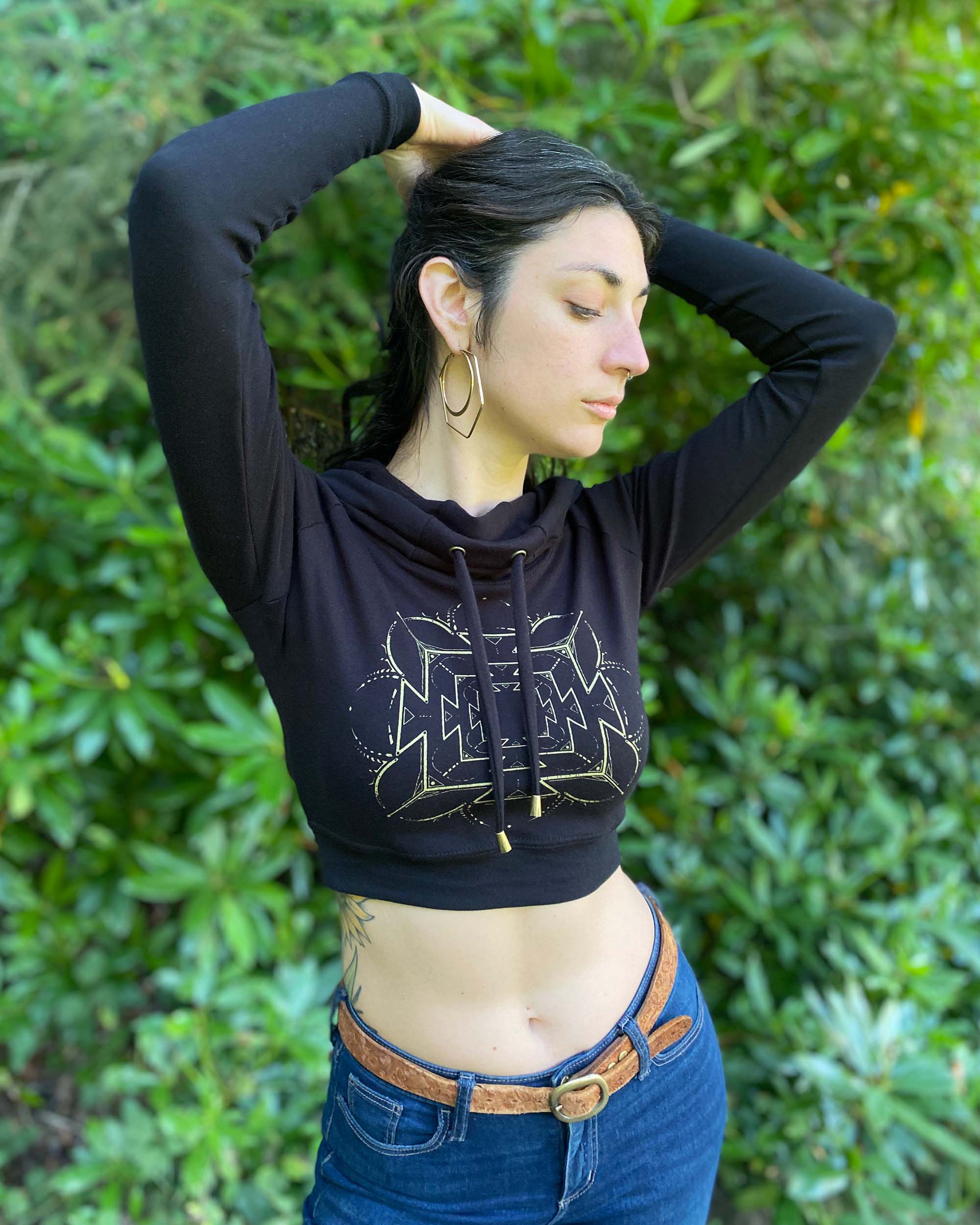 A woman wearing an organic cotton black crop hoodie with a gold print, cowl hood and long sleeves, front view.