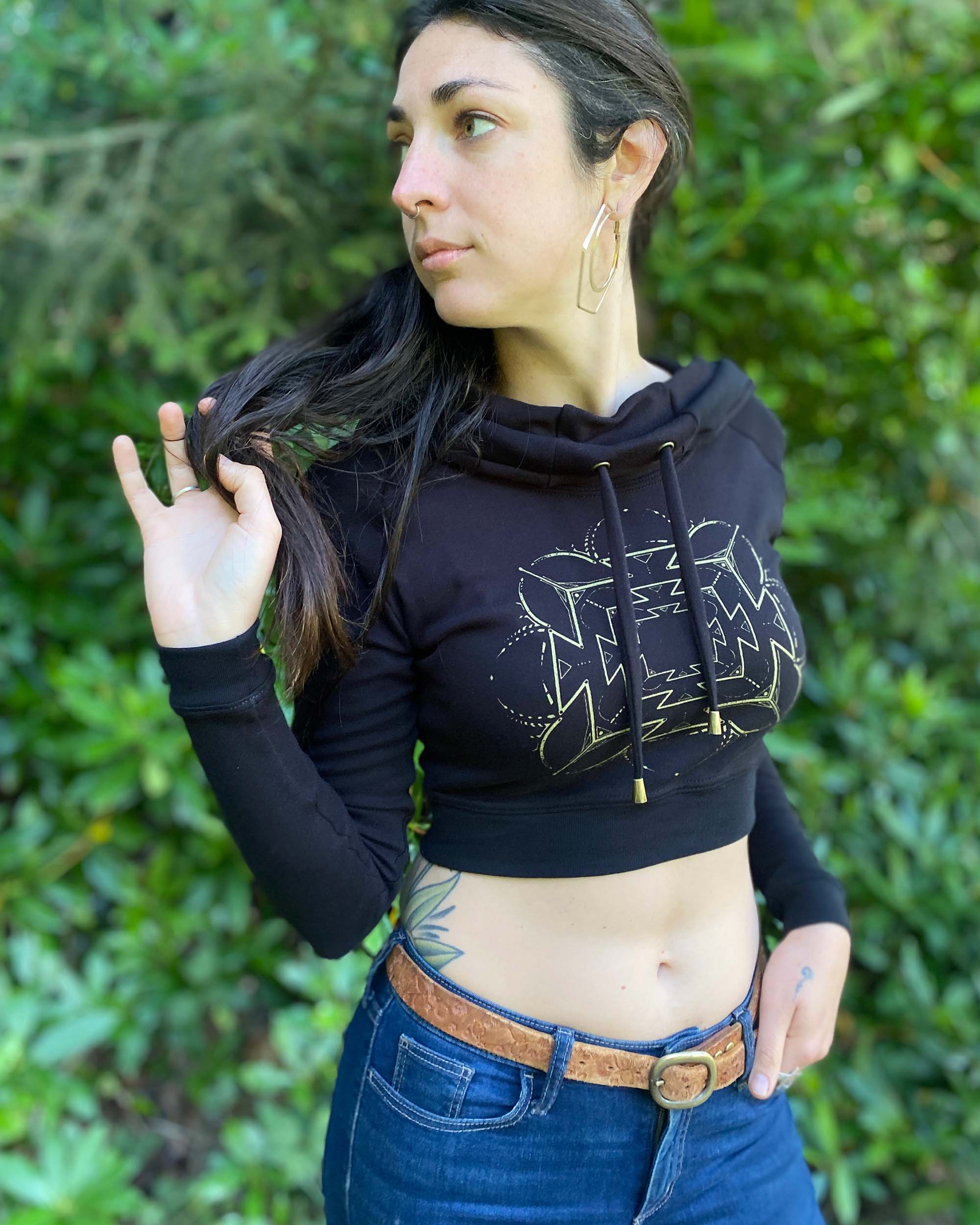 A woman wearing an organic cotton black crop hoodie with a gold print, cowl hood and long sleeves, front right side view.