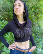 A woman wearing an organic cotton black crop hoodie with a gold print, worn with cowl hood up, close up front view.