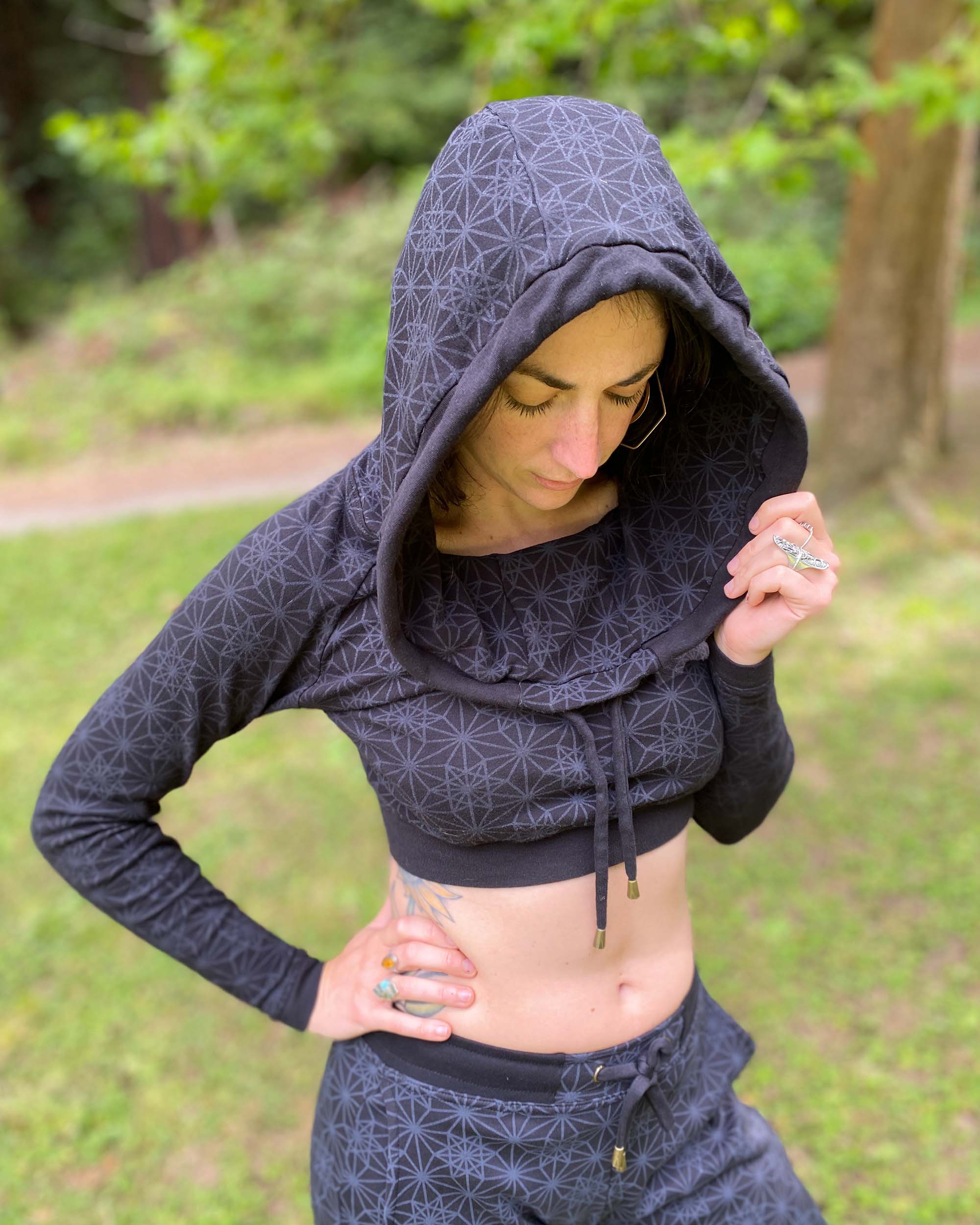 A woman wearing an organic cotton black crop hoodie with geometric print, with matching joggers, worn with hood on, front view in nature.