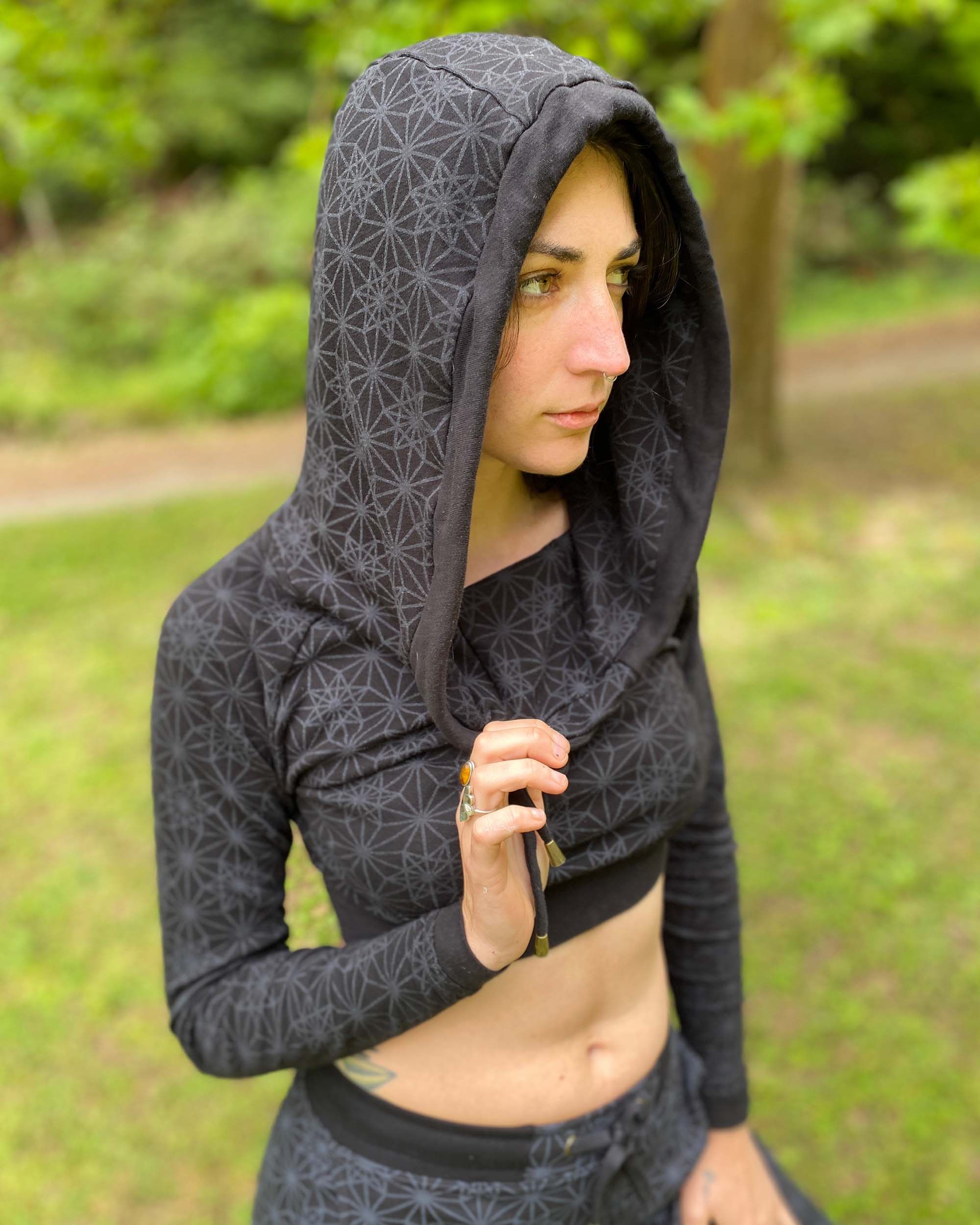 A woman wearing an organic cotton black crop hoodie with geometric print, with matching joggers, worn with hood on, side view in nature.