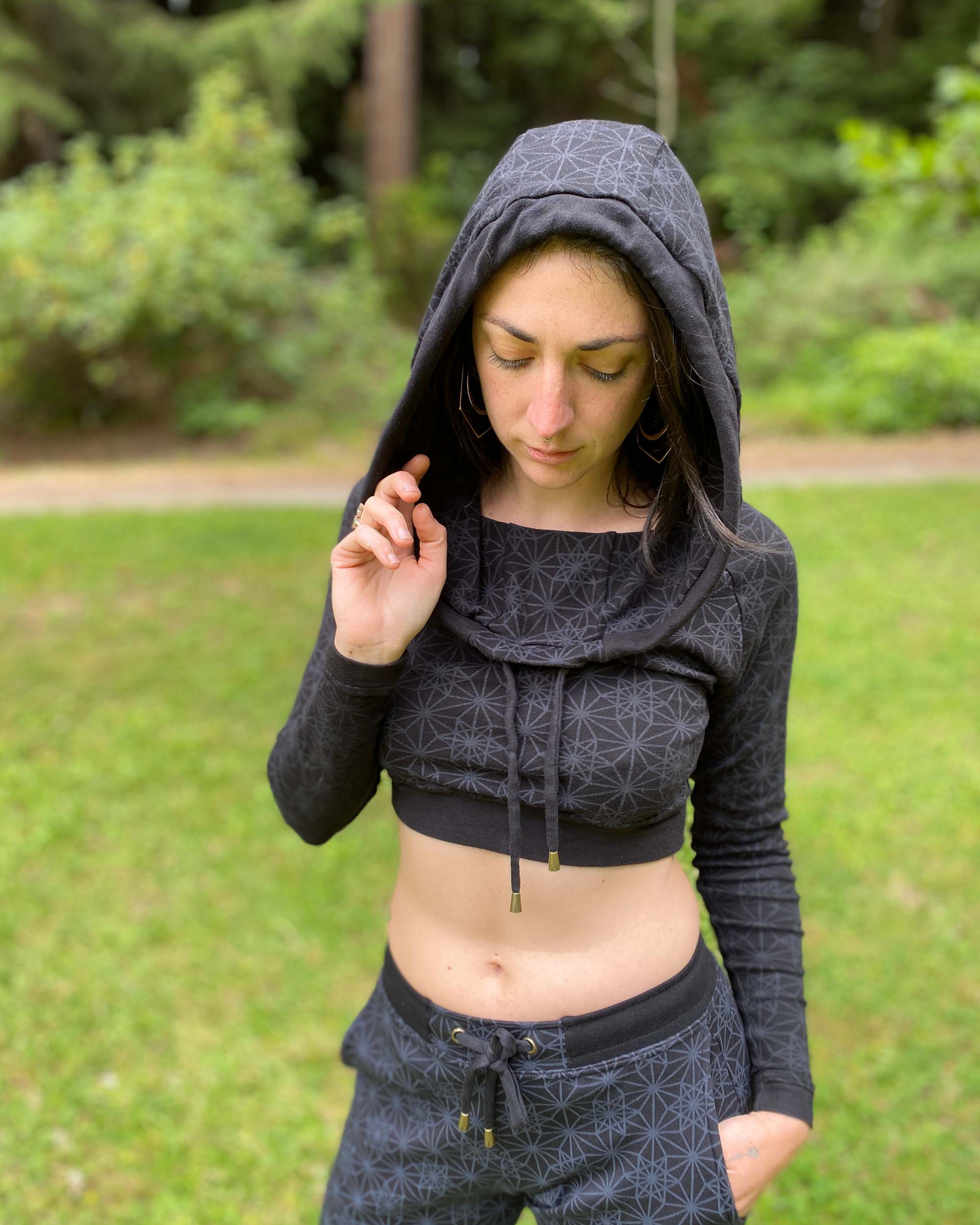 A woman wearing an organic cotton black crop hoodie with geometric printed fabric, with matching joggers, worn with hood on, front view in nature.