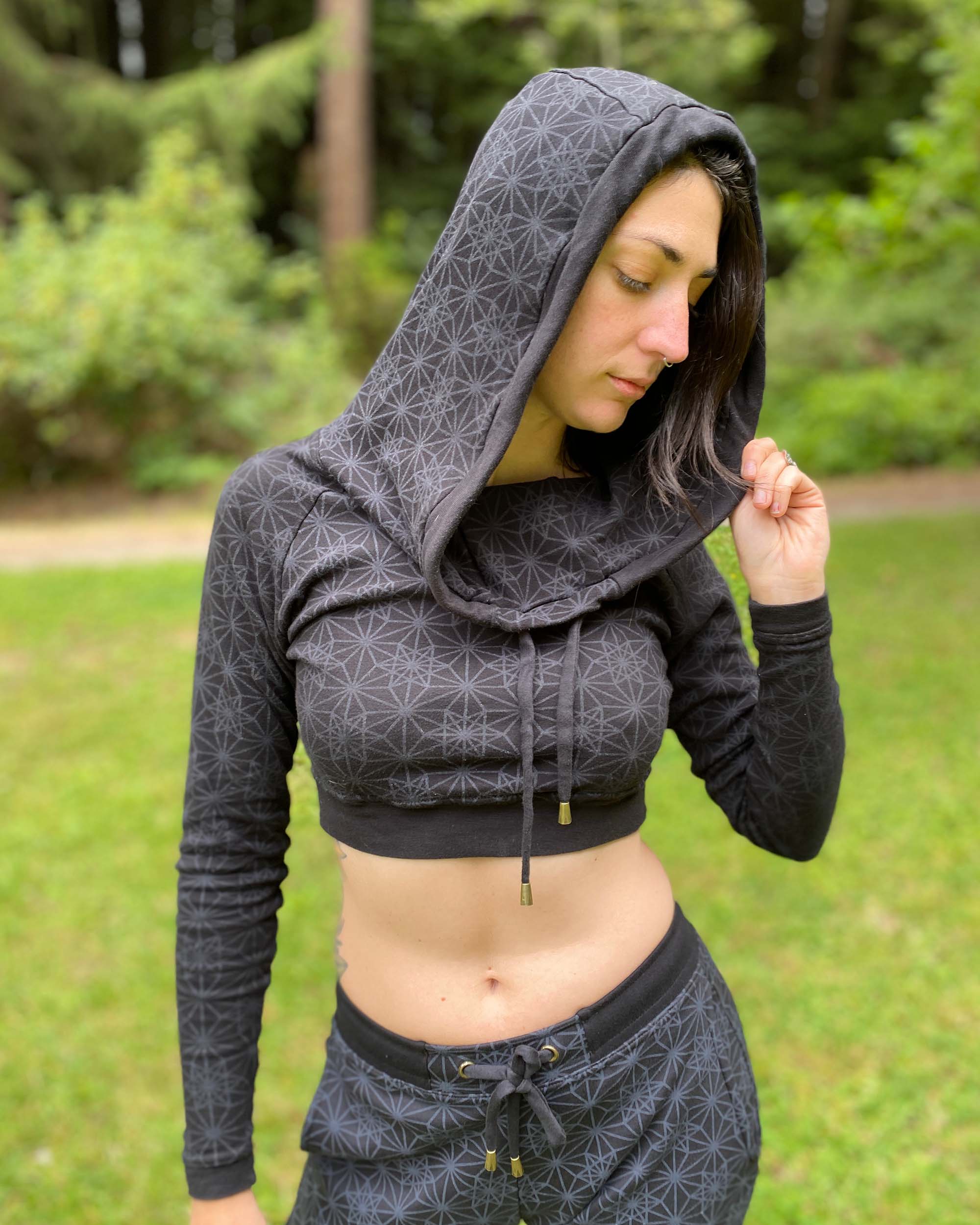 A woman wearing an organic cotton black crop hoodie with geometric print, cowl hood and long sleeves, worn with hood on, side view in nature.