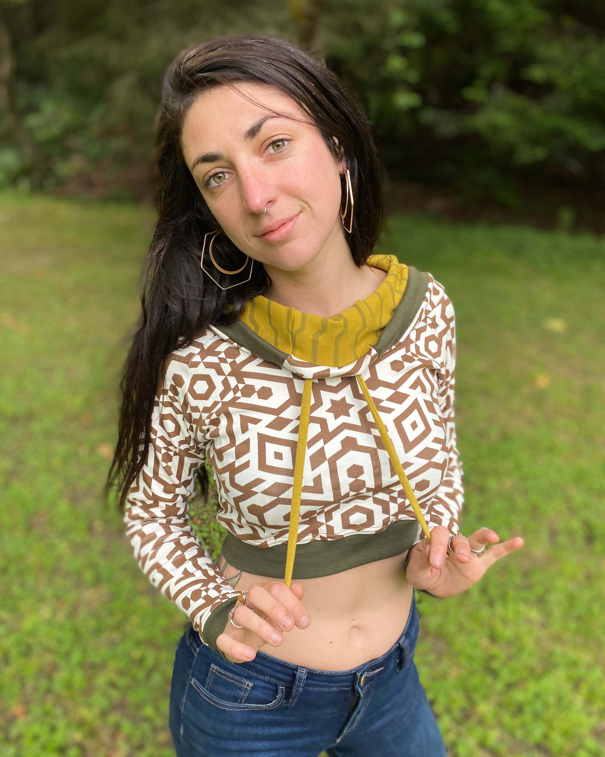 A woman wearing an organic cotton undyed crop hoodie with brown geometric printed fabric and green accents, front view in nature.