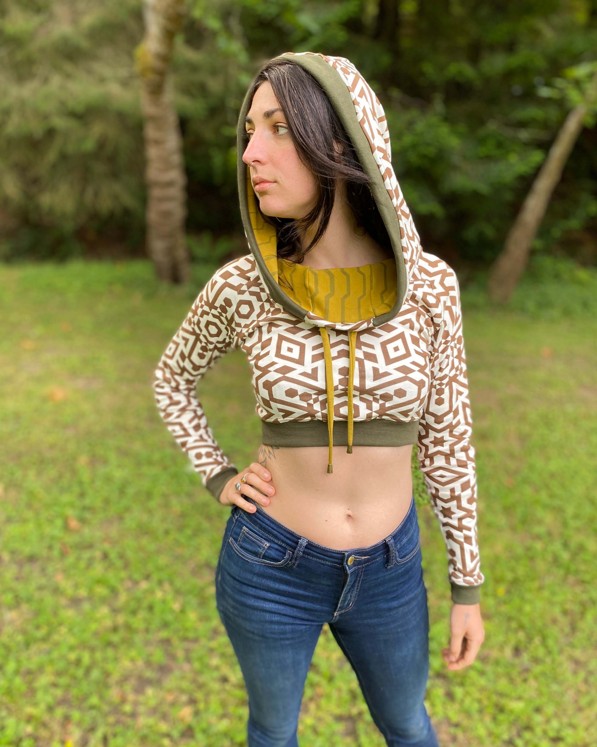 A woman wearing an organic cotton undyed crop hoodie with brown geometric printed fabric and green accents, worn with hood up, side view.