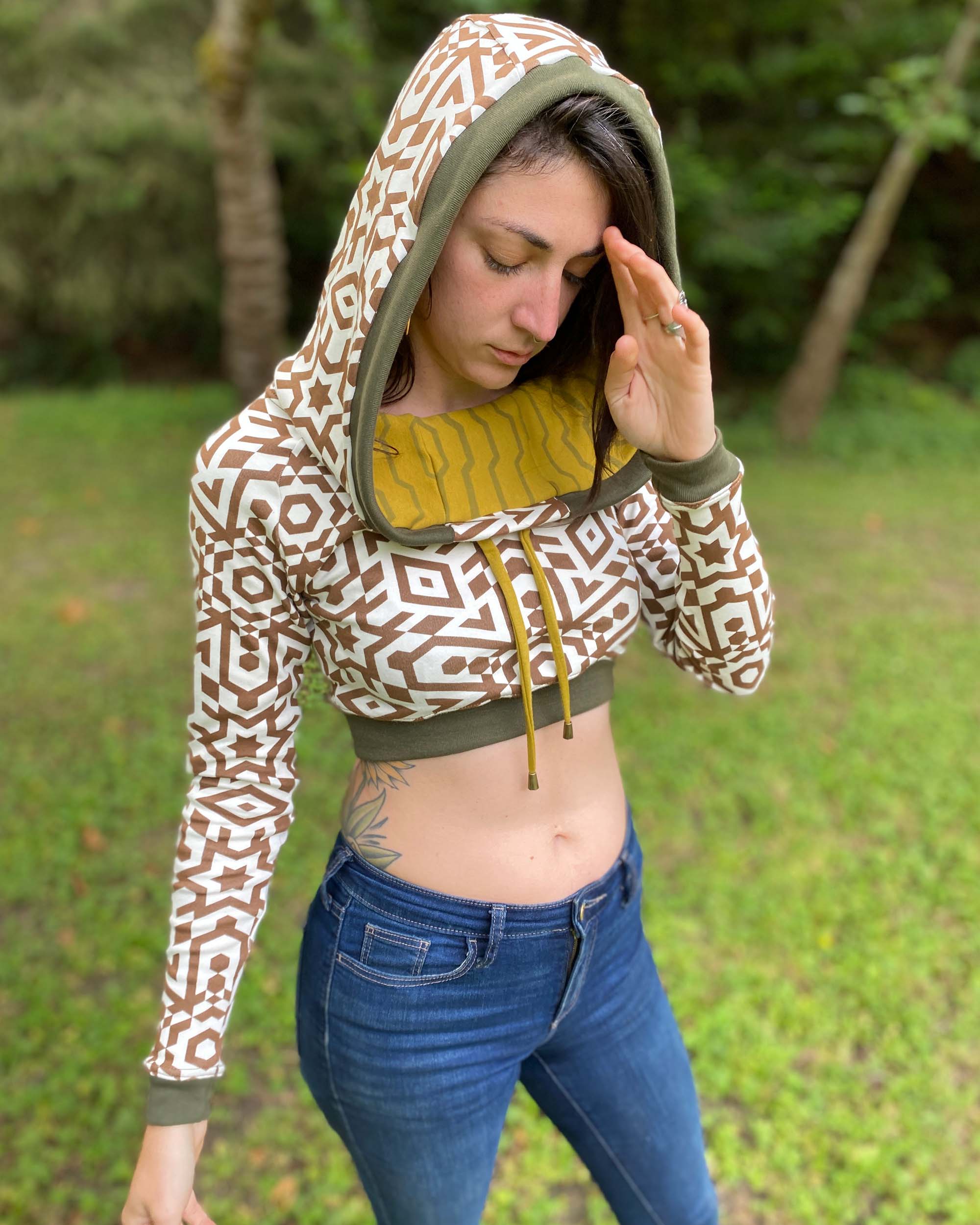 A woman wearing an organic cotton undyed crop hoodie with brown geometric printed fabric and green accents, worn with hood up, right side view.