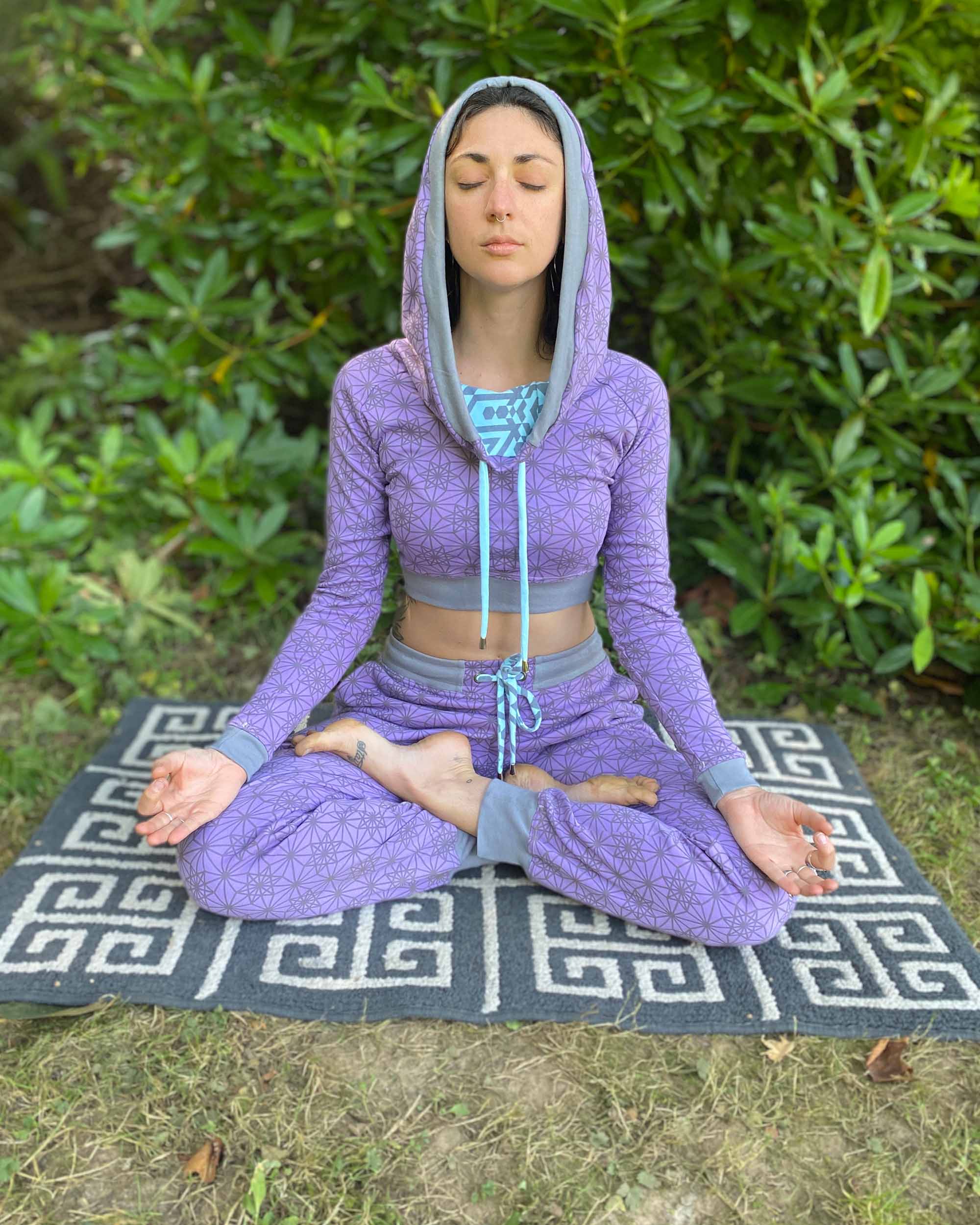 A woman meditating in nature, wearing an organic cotton lavender crop hoodie with geometric print, cowl hood and long sleeves.