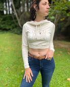 A woman wearing an organic cotton undyed crop hoodie with a gold print, cowl hood and long sleeves, front view in an outdoor setting.