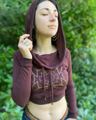 A woman wearing an organic cotton reddish brown crop hoodie with a gold print, cowl hood and long sleeves, worn with hood up, front view.