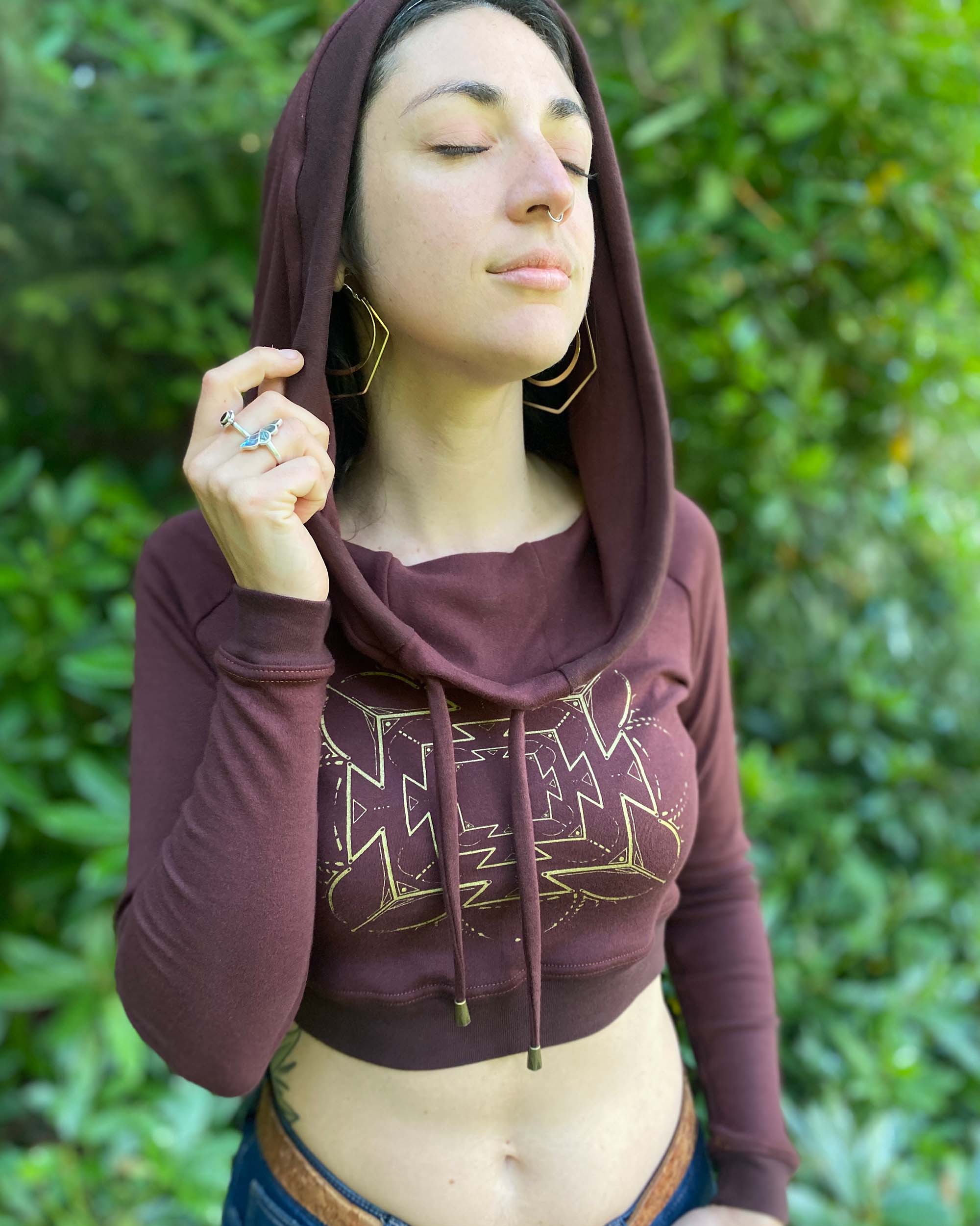 A woman wearing an organic cotton reddish brown crop hoodie with a gold print, cowl hood and long sleeves, worn with hood up, front view.