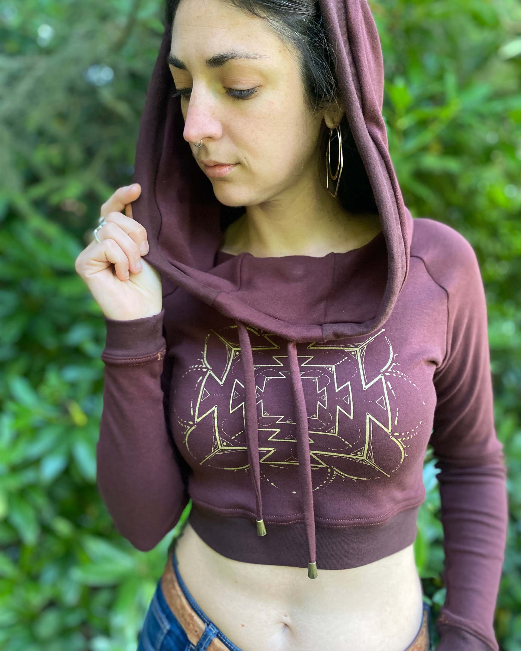 A woman wearing an organic cotton reddish brown crop hoodie with a gold print, cowl hood and long sleeves, close up view.