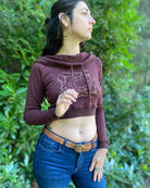 A woman wearing an organic cotton reddish brown crop hoodie with a gold print, cowl hood and long sleeves, right side view.