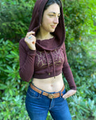 A woman wearing an organic cotton reddish brown crop hoodie with a gold print, cowl hood and long sleeves, hood up, front view.
