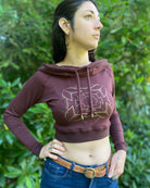 A woman wearing an organic cotton reddish brown crop hoodie with a gold print, front view.