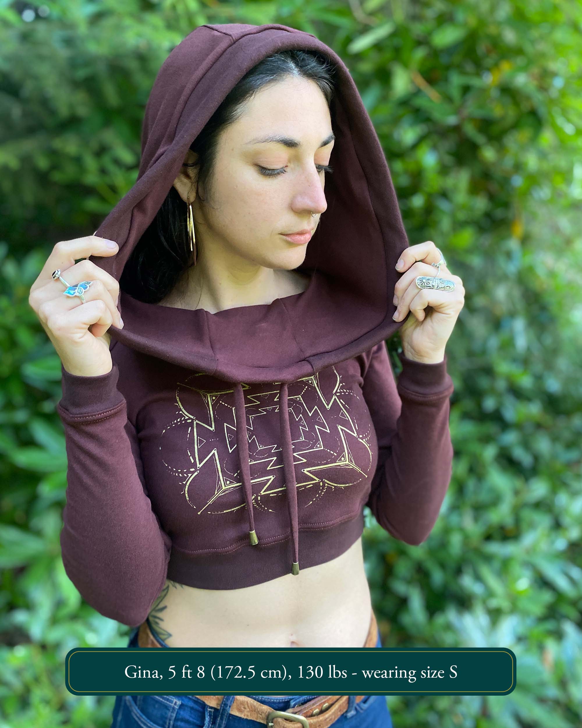 A woman wearing an organic cotton reddish brown crop hoodie with a gold print, cowl hood and long sleeves, front view.