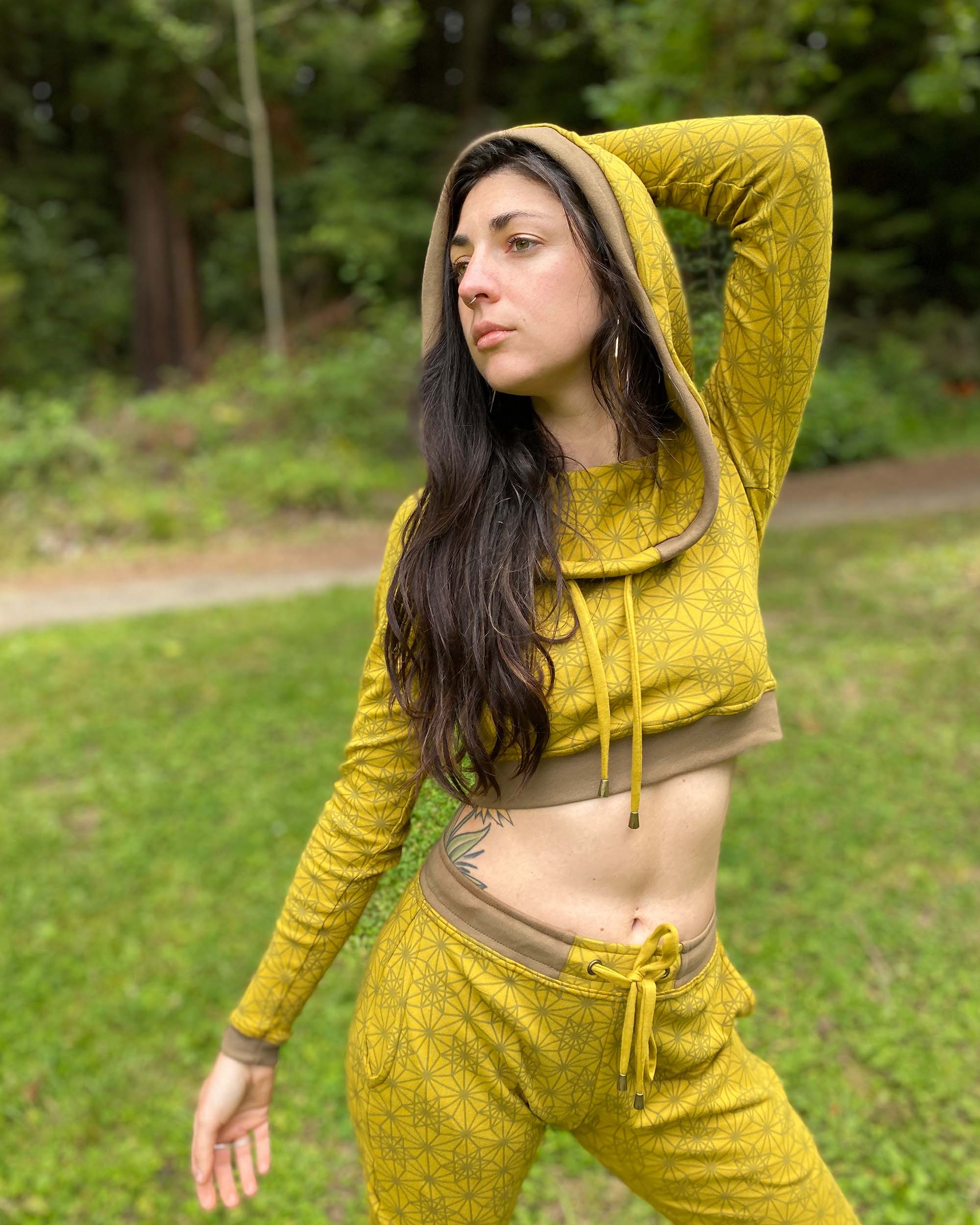 A woman wearing an organic cotton yellow crop hoodie with geometric print, cowl neck and long sleeves, front view in a outdoor setting.