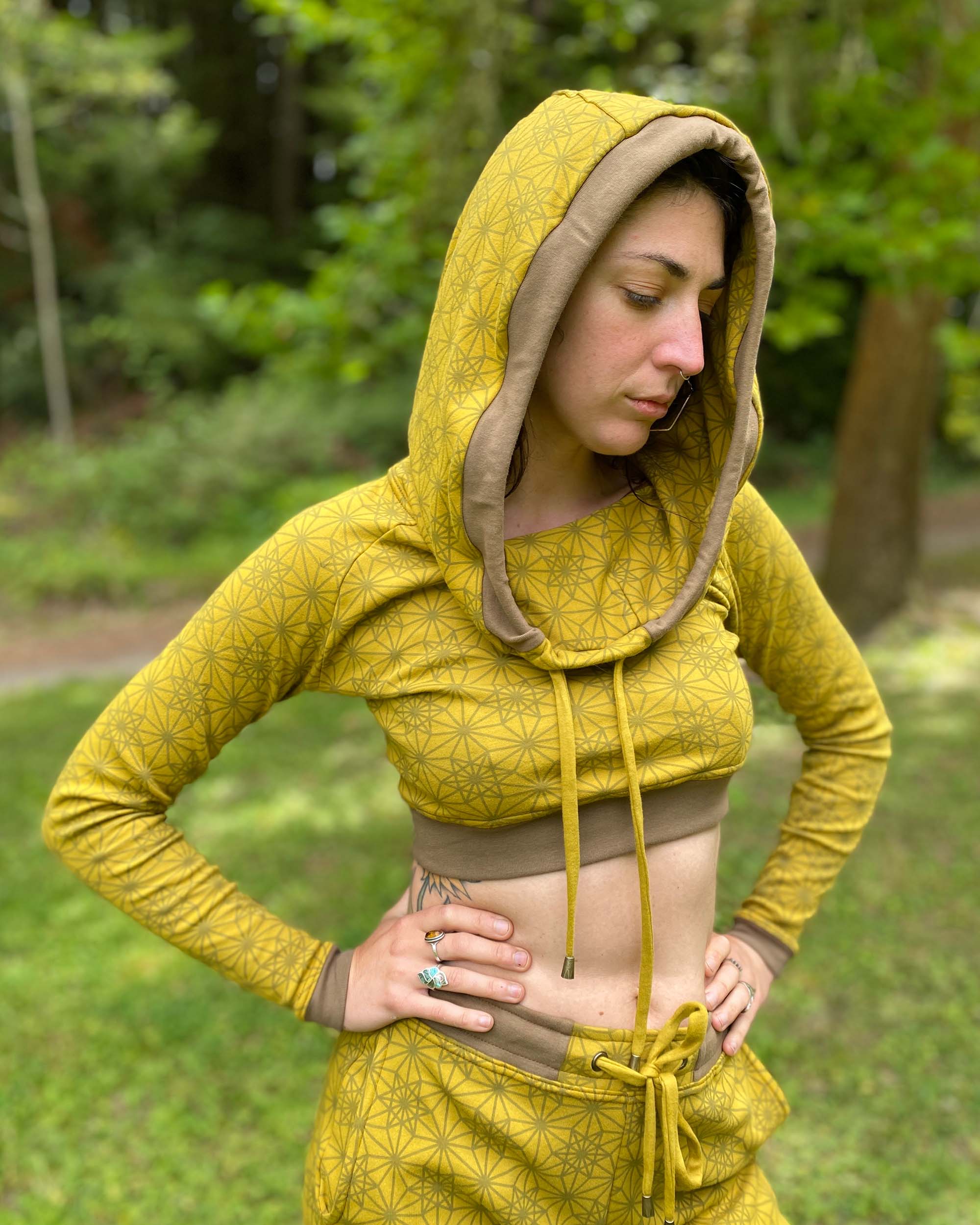 A woman wearing an organic cotton yellow crop hoodie with geometric print, with matching joggers, front right side view in a outdoor setting.