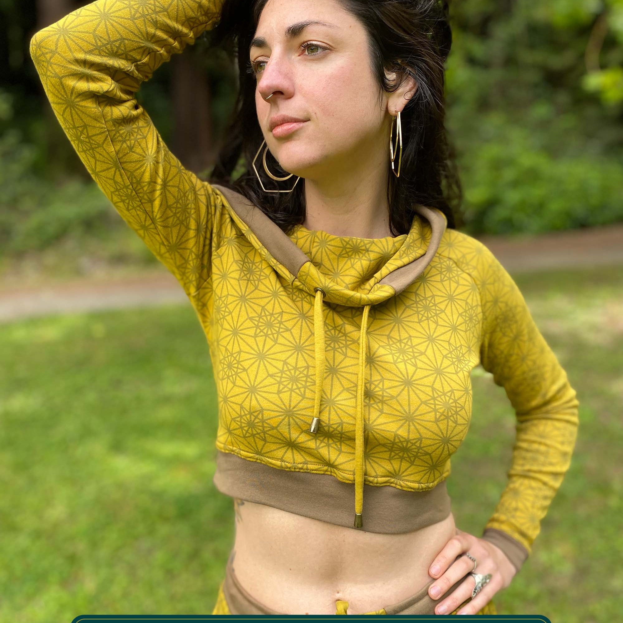 A woman wearing an organic cotton crop hoodie, yellow with geometric print, cowl neck and long sleeves, front view in a outdoor setting.