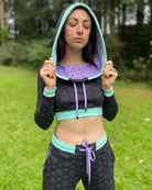 A woman wearing an organic cotton black crop hoodie with geometric print and purple accents, cowl hood and long sleeves, worn with hood on.