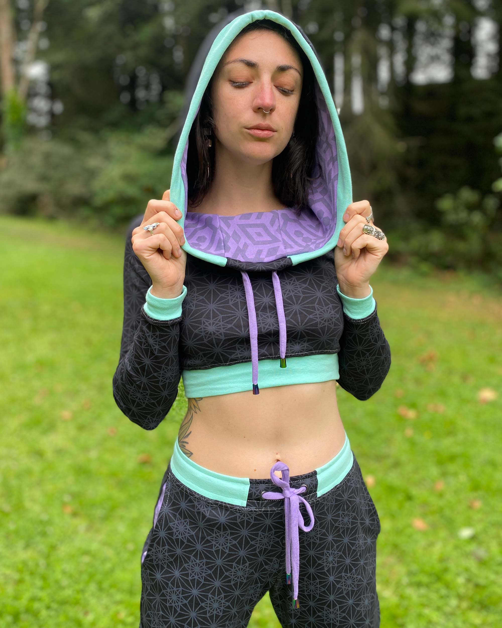 A woman wearing an organic cotton black crop hoodie with geometric print and purple accents, cowl hood and long sleeves, worn with hood on.