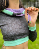 A woman wearing an organic cotton black crop hoodie with geometric print and purple accents, cowl hood and long sleeves, close up view of anodized drawcord details.