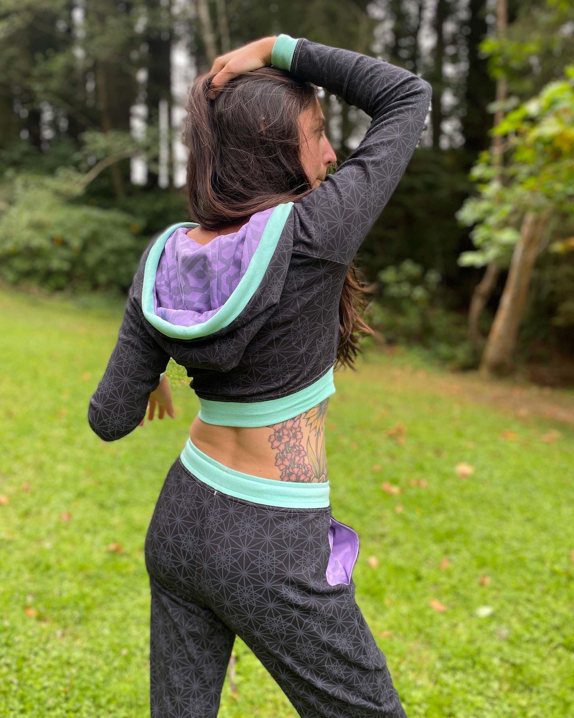 A woman wearing an organic cotton black crop hoodie with geometric print and purple accents with matching joggers, back view.