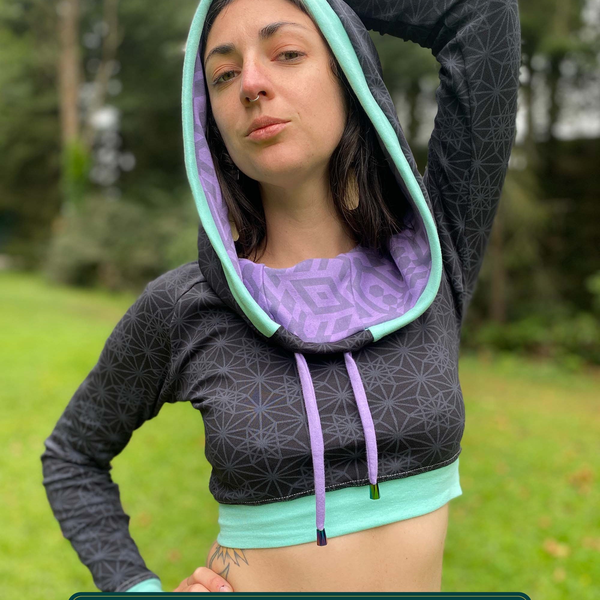 A woman wearing an organic cotton black crop hoodie with geometric print and purple accents, cowl hood and long sleeves, front view with hood up.