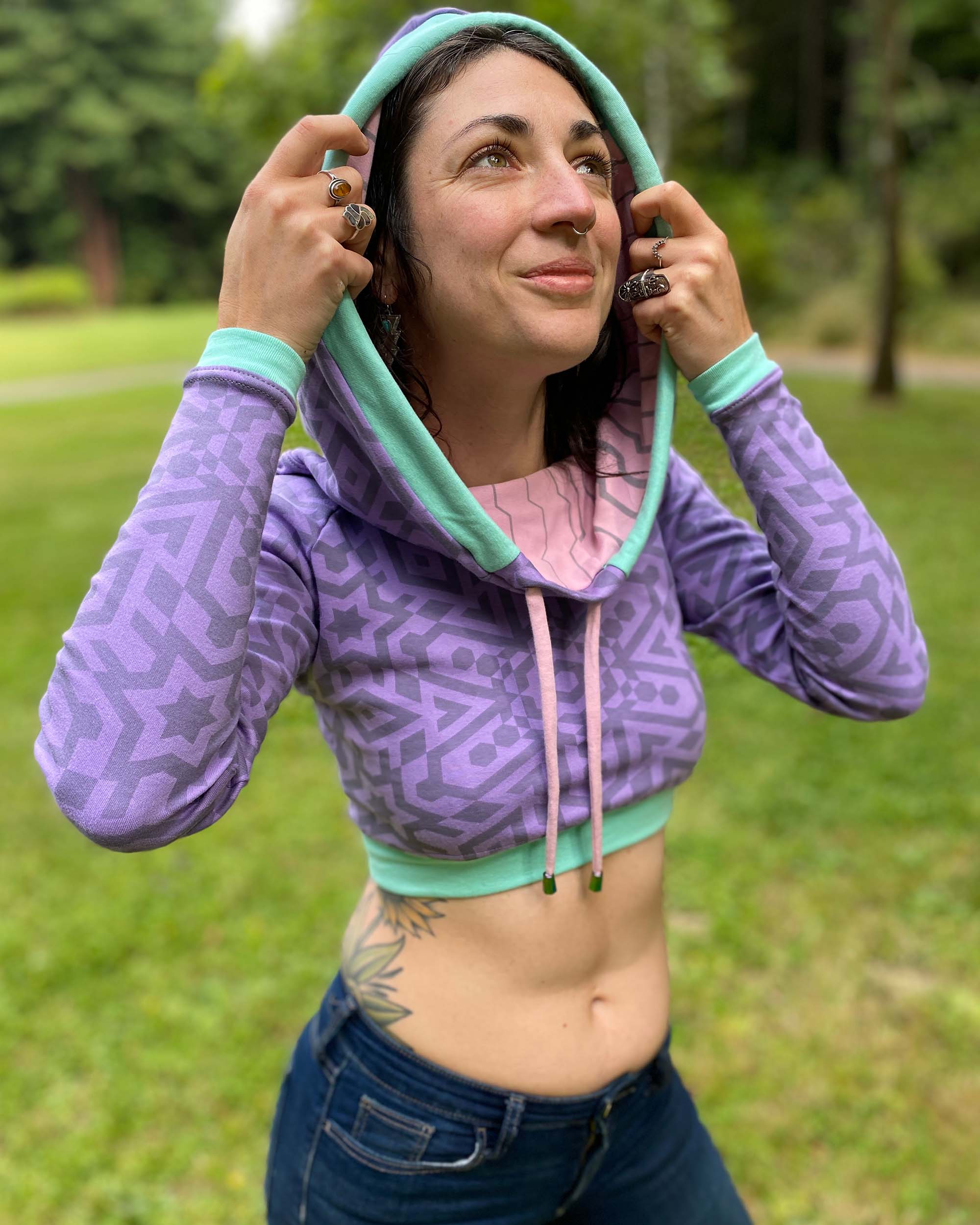 A woman wearing an organic cotton lavender crop hoodie with geometric print and pink accents, worn with cowl hood up, front side view.