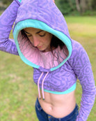 A woman wearing an organic cotton lavender crop hoodie with geometric print and pink accents, worn with cowl hood up, left side view.