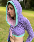 A woman wearing an organic cotton lavender crop hoodie with geometric print and pink accents, worn with cowl hood up, left side view.
